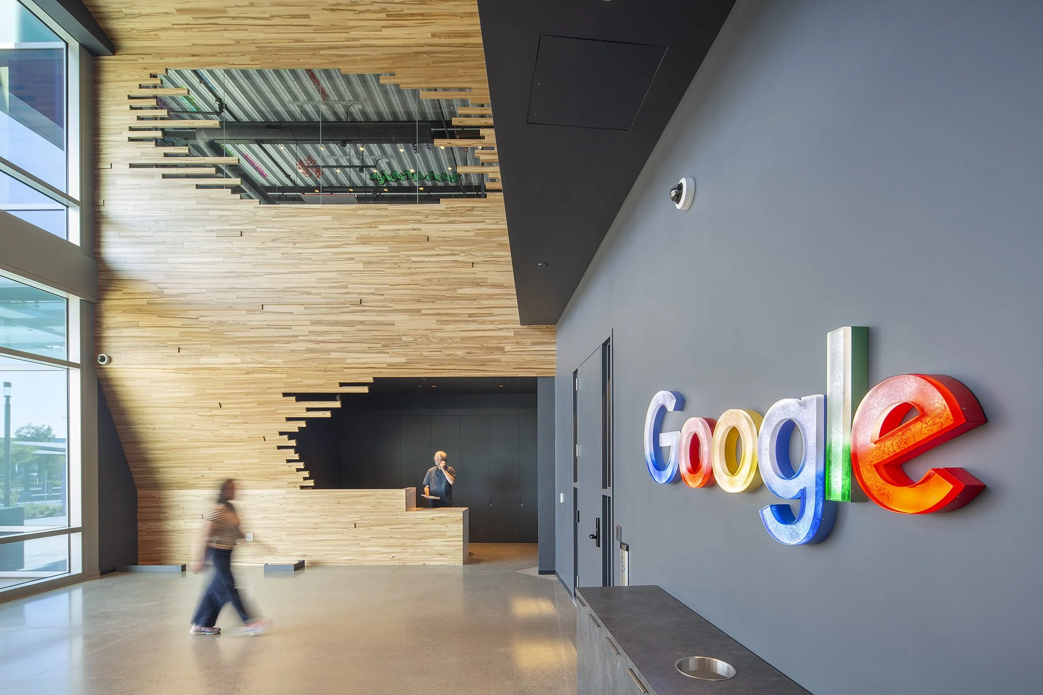 Interior of a Google office lobby with a colorful illuminated Google logo, wooden wall design, and large windows. Two blurred people are walking inside.