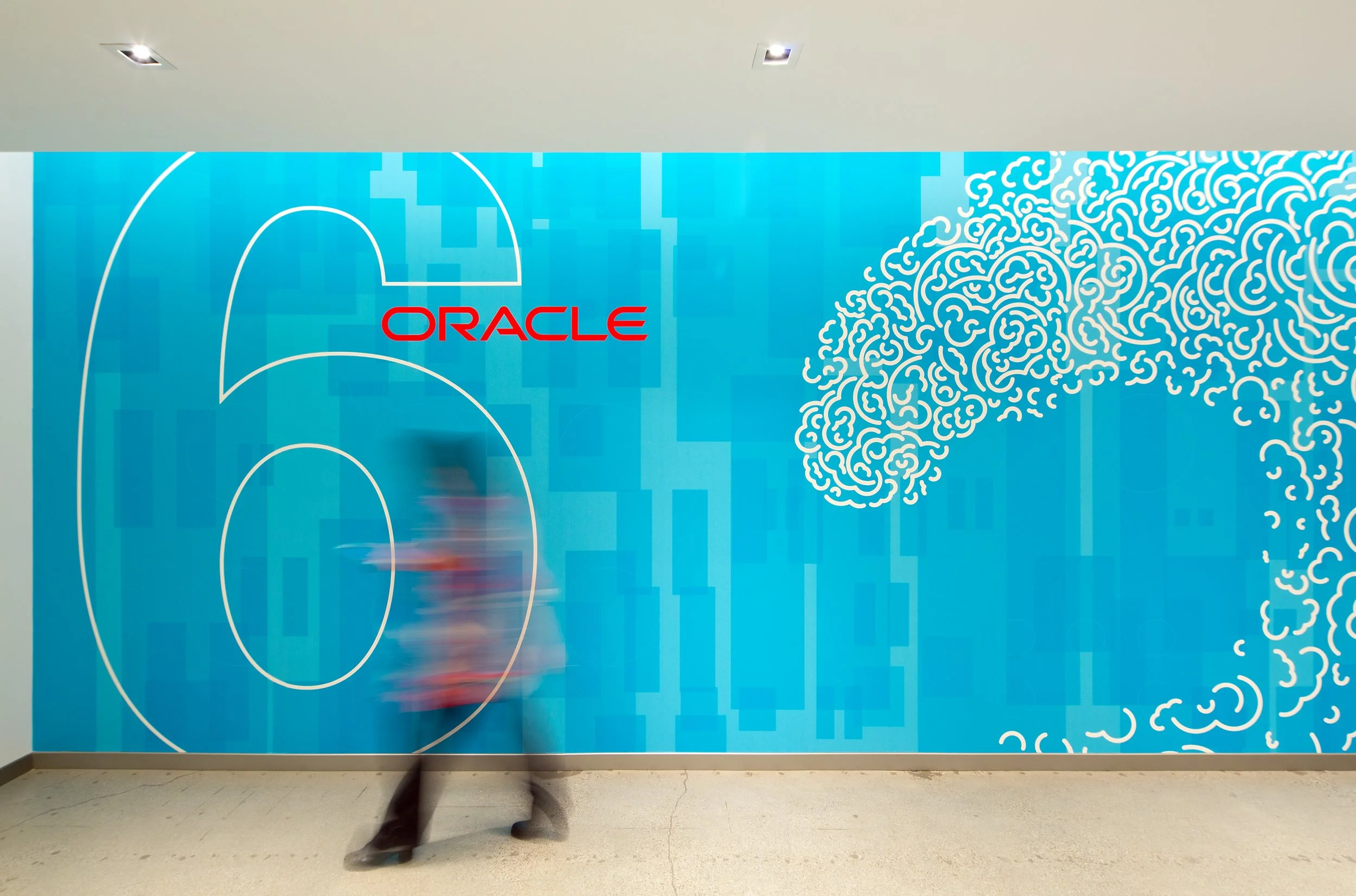 Blue wall with a white and red Oracle logo, abstract white line patterns, and a person walking in front of it, captured with motion blur.
