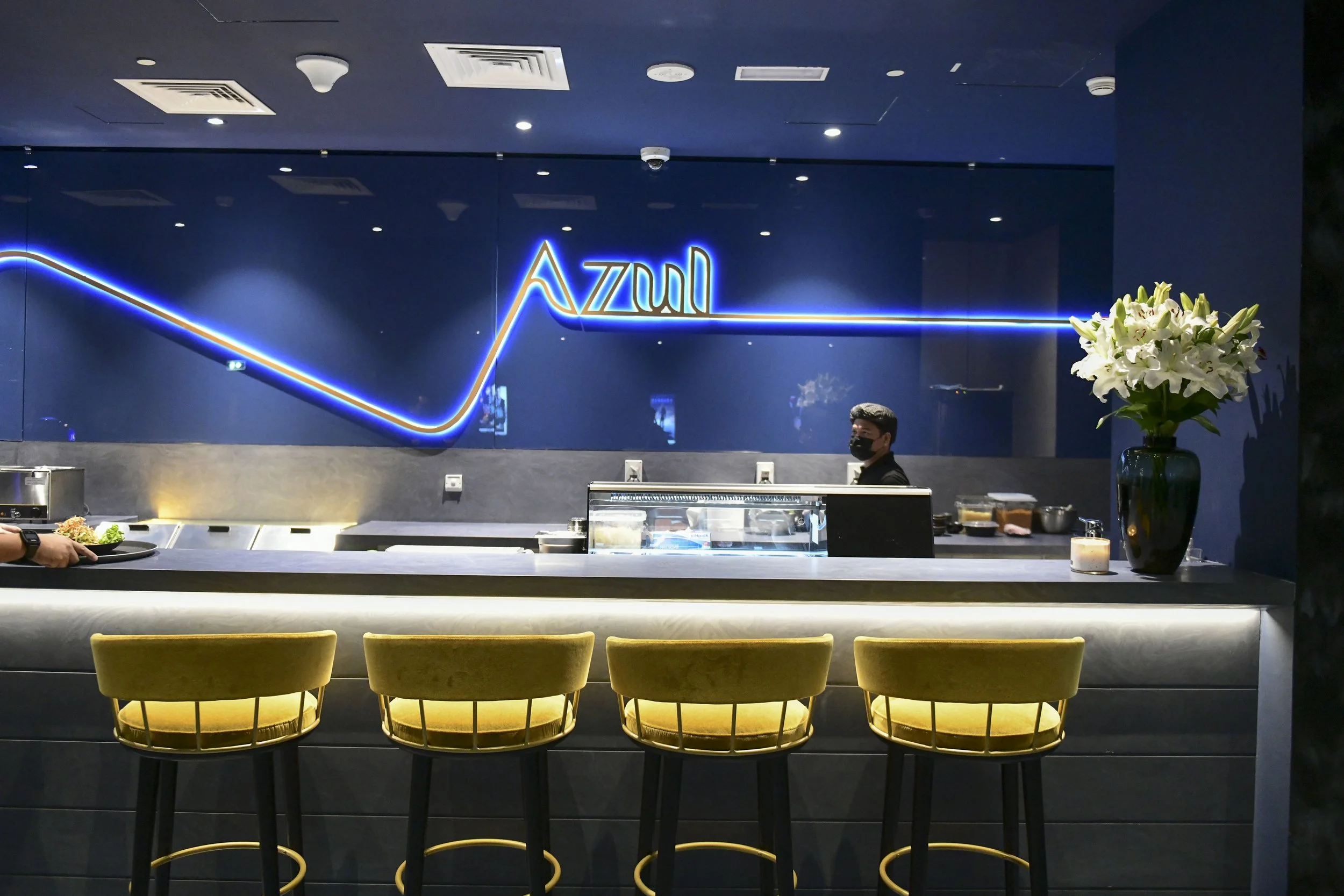 Interior of a modern restaurant with a counter, yellow chairs, a neon sign that reads 'Azuhl,' a large flower bouquet on the right side, and a person behind the counter wearing a face mask.