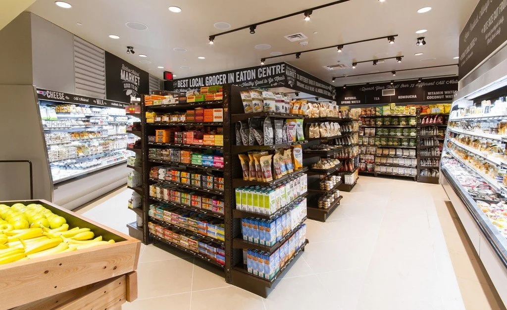 Interior view of a grocery store aisle with shelves stocked with packaged food items, a display of bananas in the foreground, and signs indicating different sections like cheese, milk, and beverages.