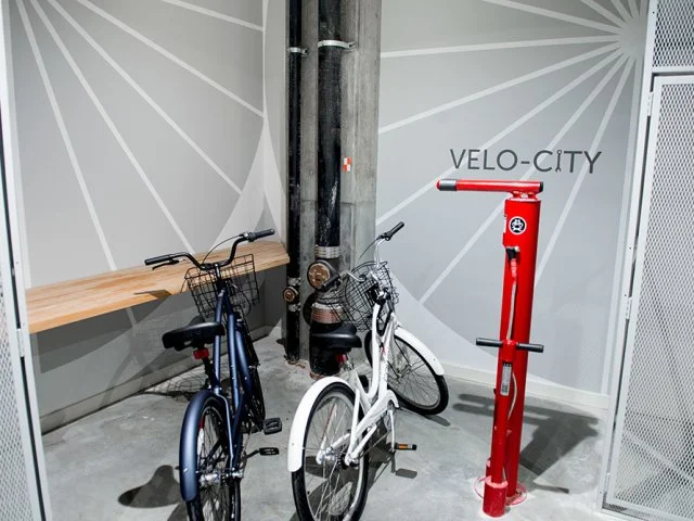 Two bicycles, one blue and one white, are parked near a wall with a sign that says 'VELO-CITY'. A red bike pump is nearby, and a wooden bench is against the wall on the left side.