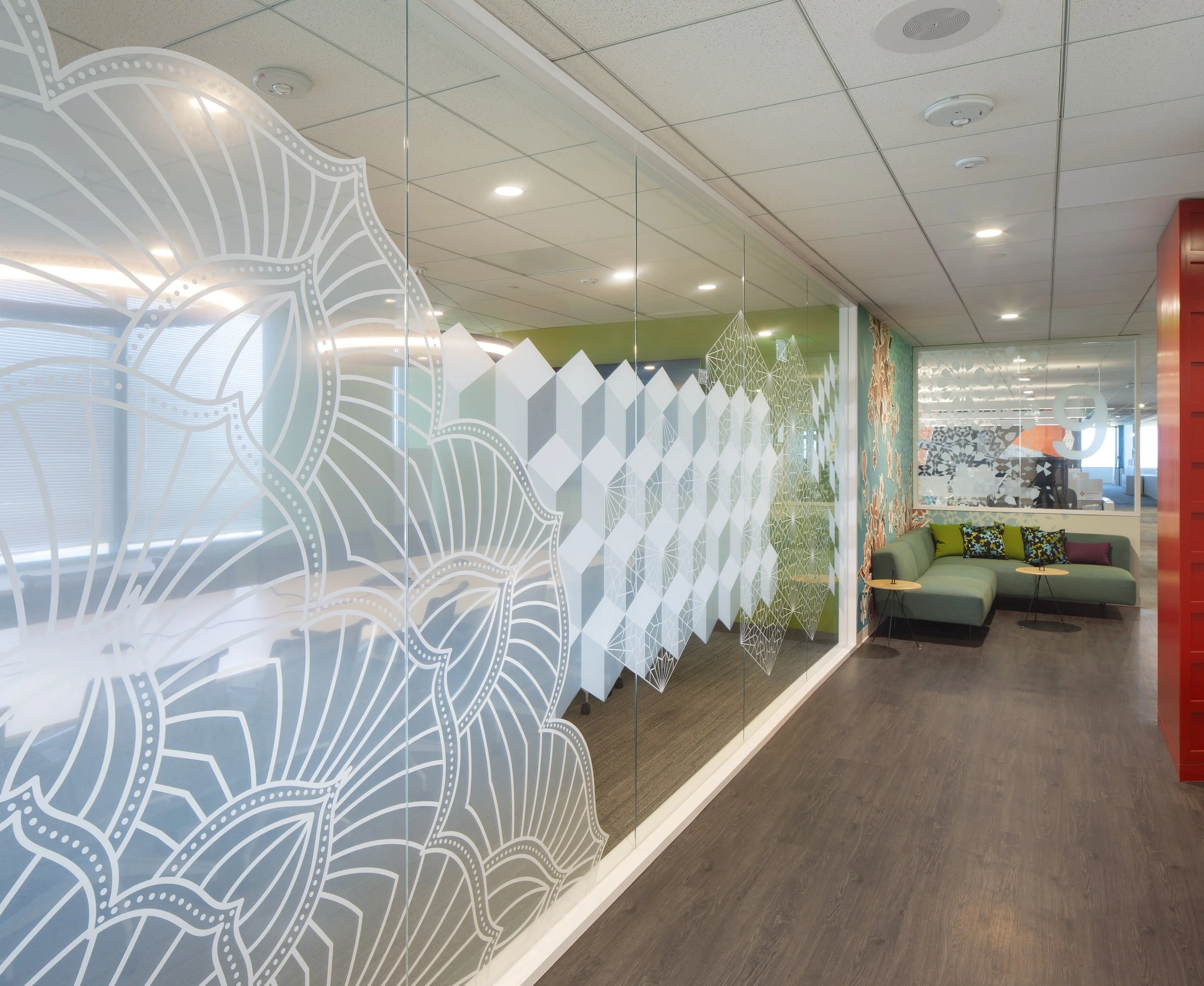 Interior of an office with glass walls featuring white geometric and floral patterns, seating area with green sofa and decorative cushions, artwork on the wall, ceiling lights, and a door with patterned glass.