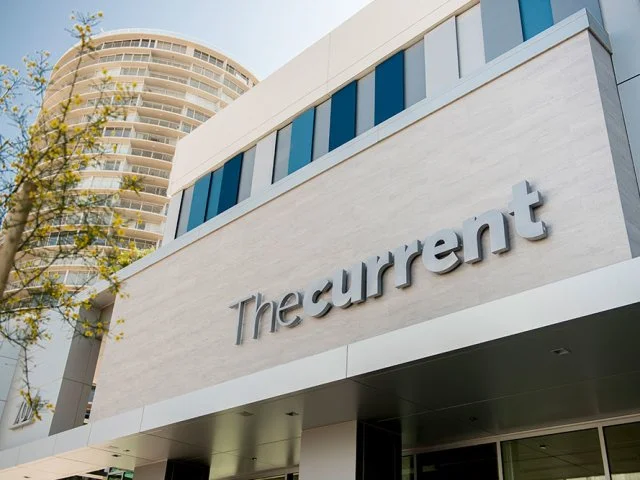 A modern building with a sign that reads 'The current' on its exterior. In the background, there is a tall, rounded high-rise apartment or office building and a tree with yellow-green leaves.