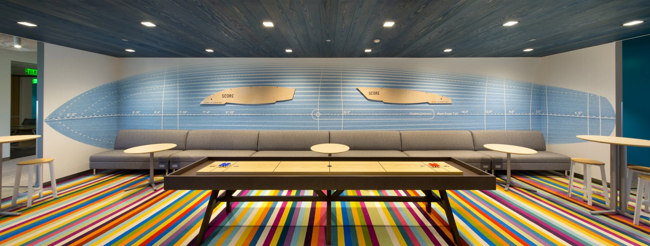 Indoor lounge with a colorful striped carpet, a large gray sectional sofa, and a shuffleboard table in the foreground. The back wall has a mural of a blue watersports track with measurement markings and scores.