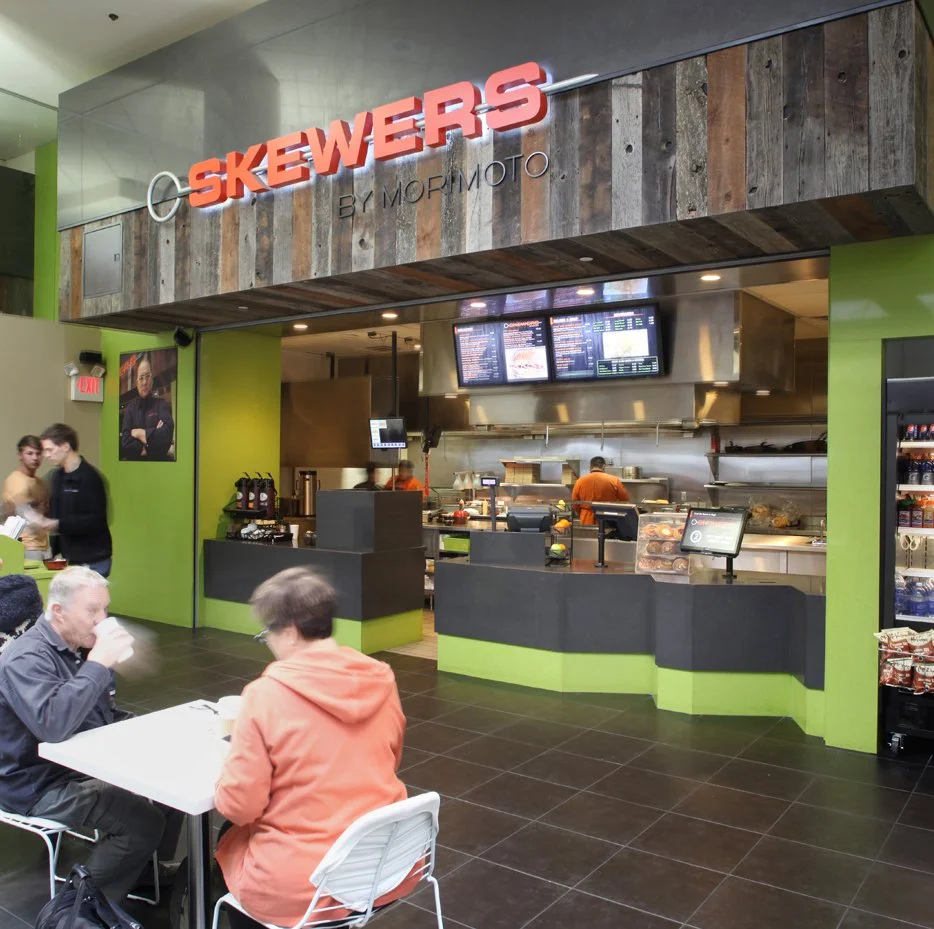 Interior of a food restaurant called Skewers by MoMoTo, with a counter for ordering food and a menu displayed on digital screens above. Customers are seated at tables, and staff are working behind the counter in the kitchen area.