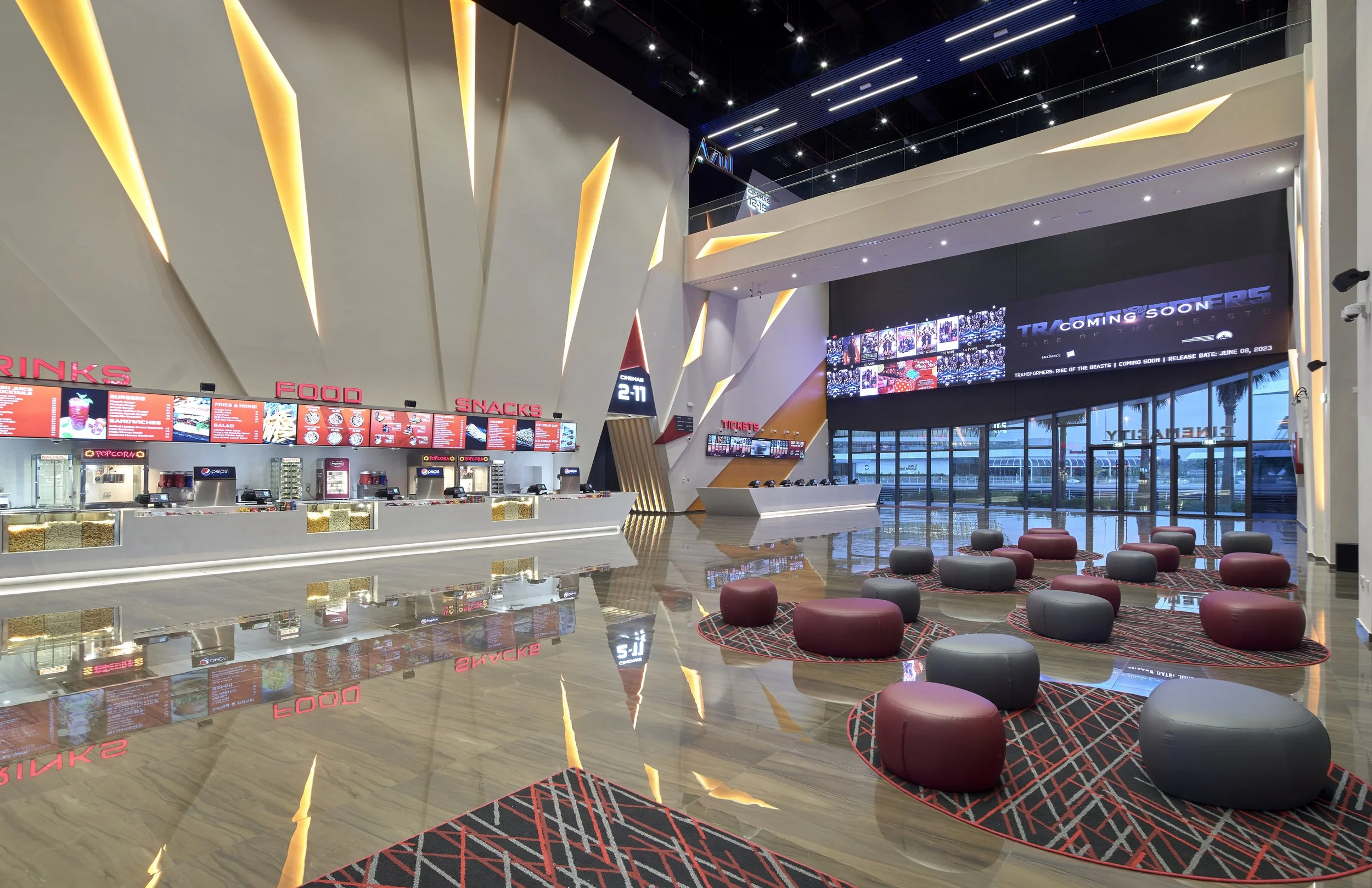 Modern movie theater lobby, digital screens displaying food and ticket information, seating area with red and gray ottomans, and large glass windows with palm trees outside.