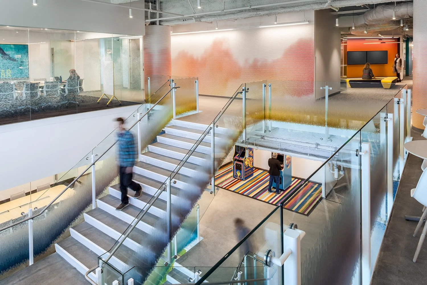 Interior of a modern office or public building with glass walls, a colorful staircase, and a hallway with workspaces and seating areas. There are people walking and sitting inside the space.