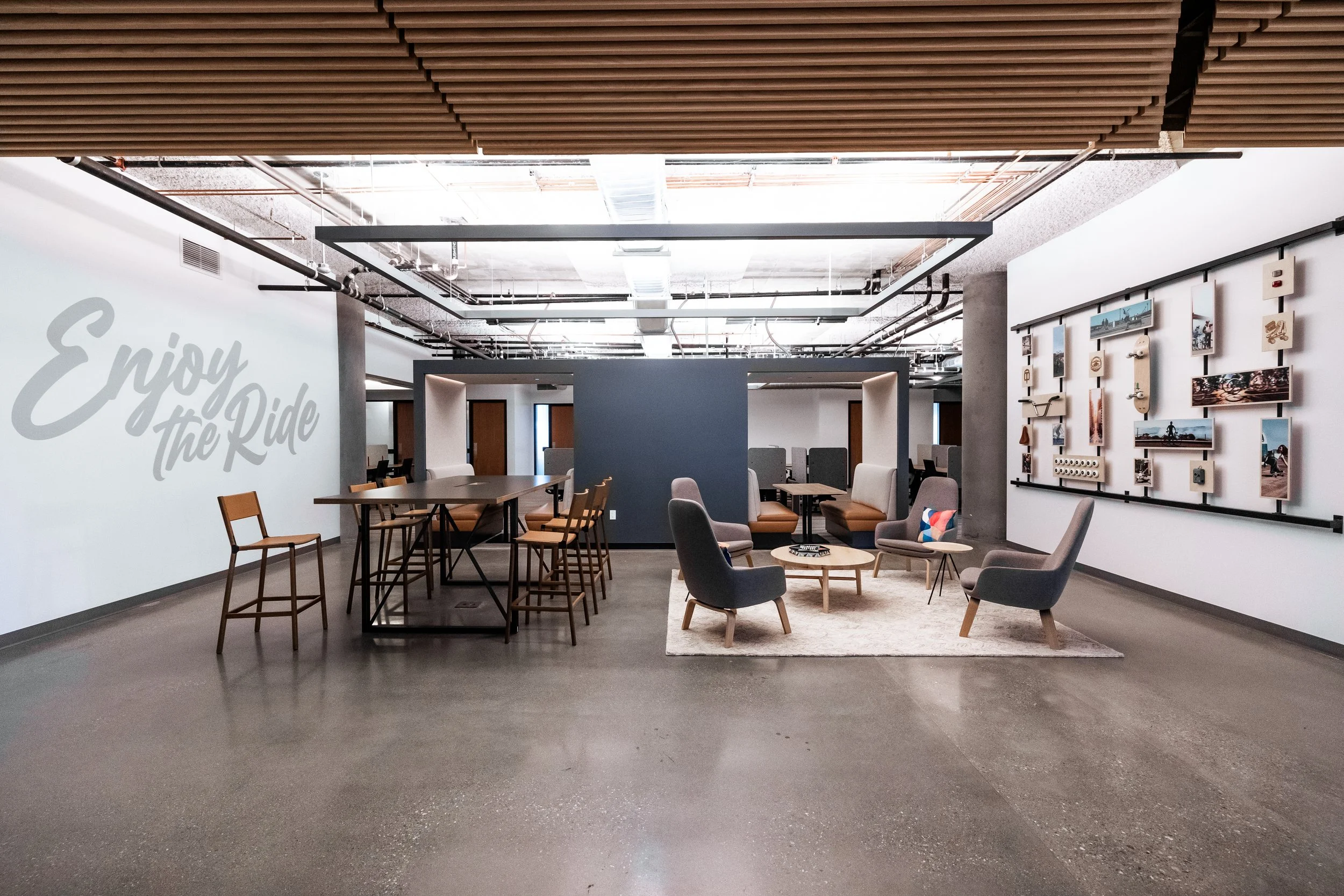 Interior of a modern lounge or common area with various seating options including chairs and sofas, wooden and fabric, a wall display with photos, and a large wall sign that reads "Enjoy the Ride". The ceiling features exposed pipes and wooden slats.