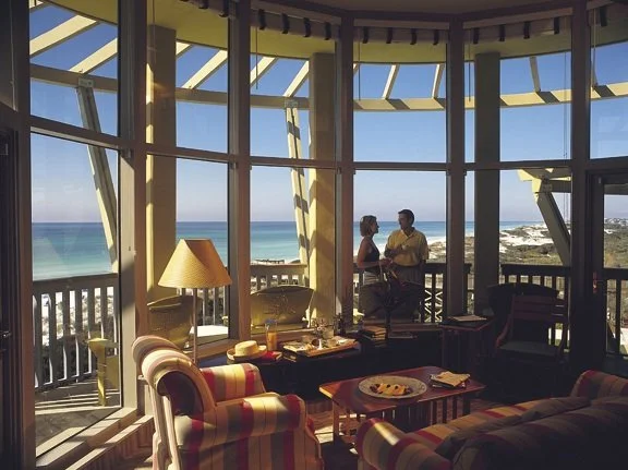 Cozy living room with large windows overlooking the ocean, featuring armchairs, a lamp, and a tabletop with snacks, with two people standing outside on a balcony.