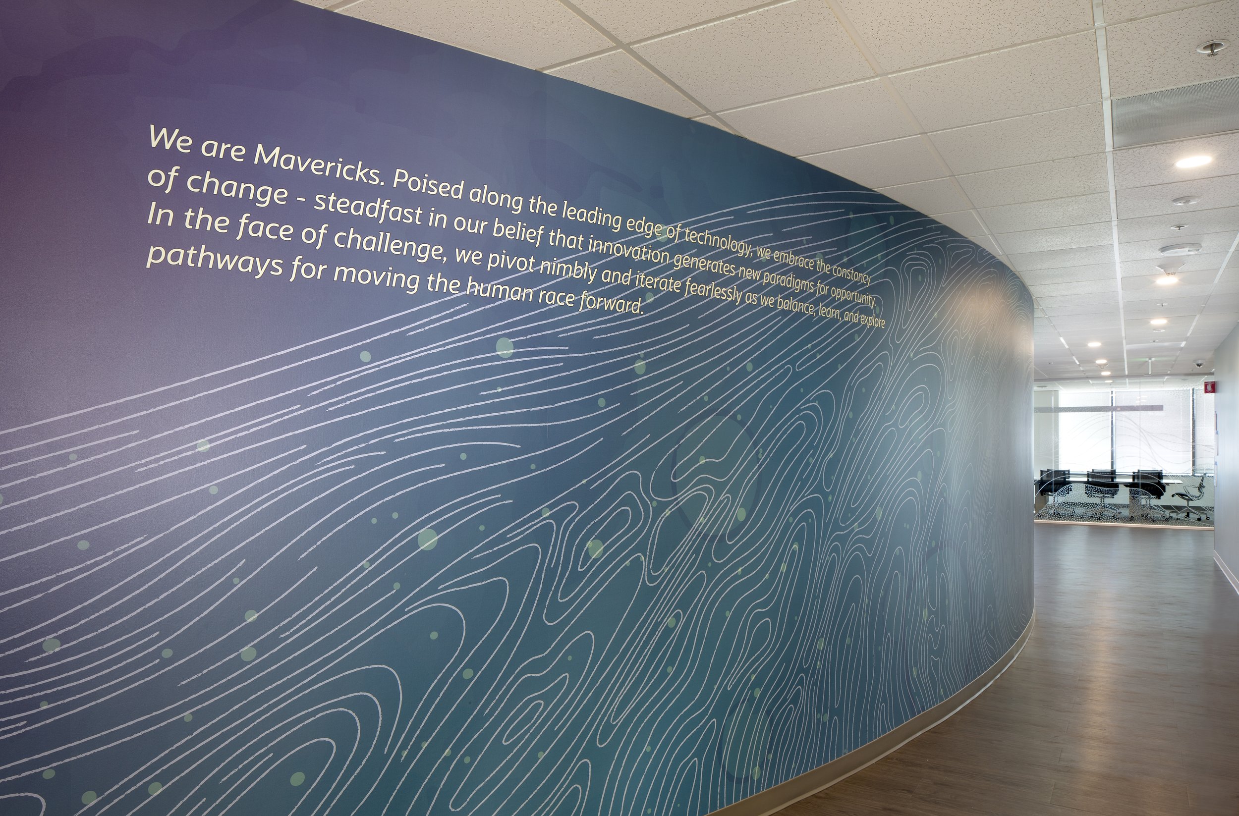 Interior view of a modern office or corporate space with a large wall display featuring a quote about innovation and change. The wall has a blue and purple abstract design with white lines and green dots, and a corridor with ceiling lights leading to