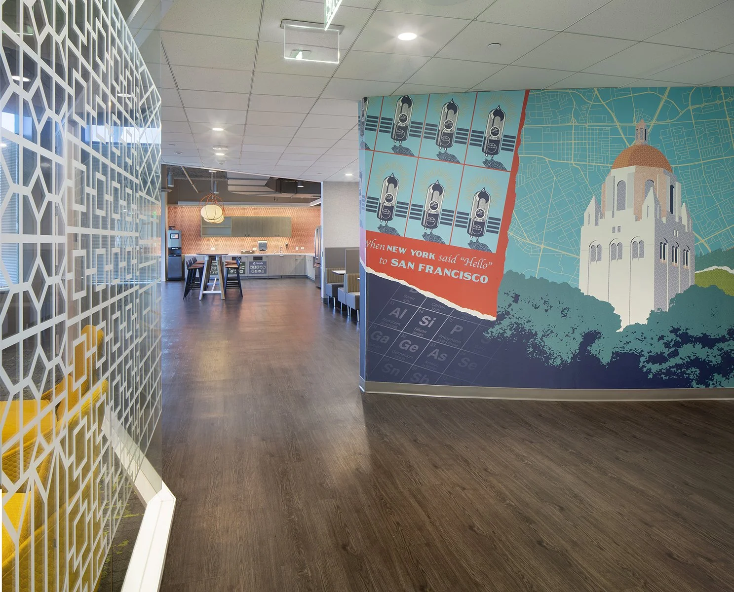 Interior view of an office or lounge area with a wall featuring a mural. The mural depicts a vintage travel poster from New York to San Francisco, with illustrations of a rocket and a cityscape. The space has wooden flooring, a seating area with armc