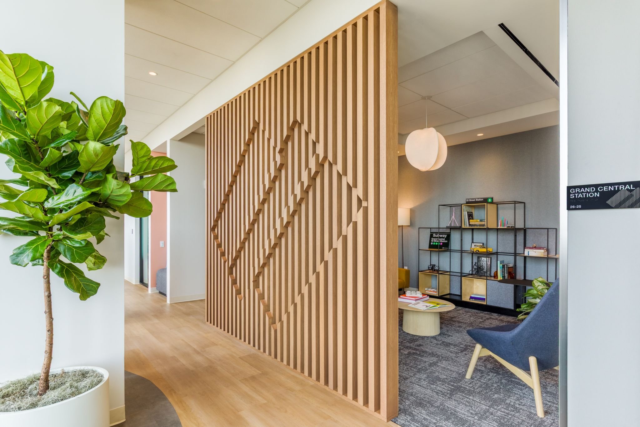Interior of a modern office or lobby with a wooden decorative divider, a potted green plant, and a bookshelf with books and decor items.
