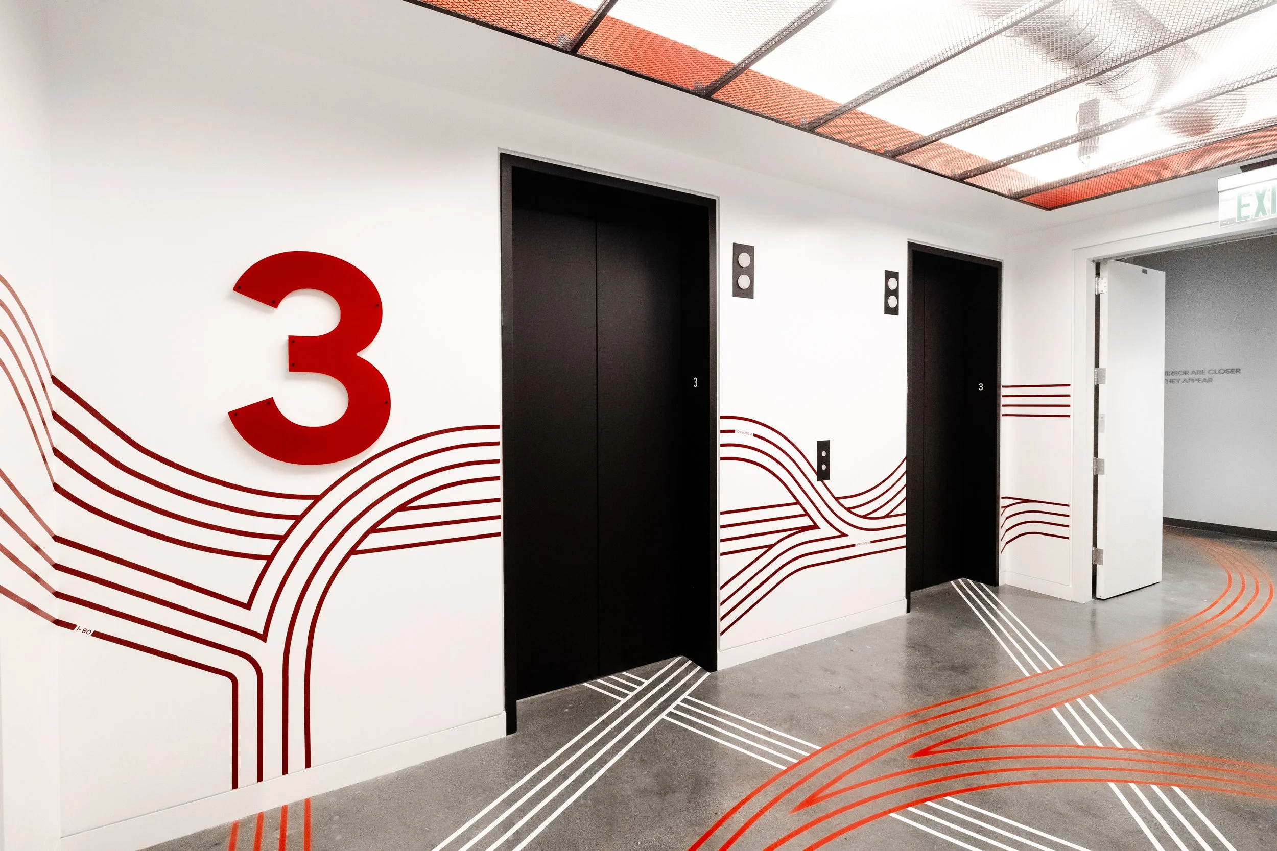 Modern interior hallway with black elevator doors, white walls decorated with red and gray line patterns, and a ceiling with red grid panels and ceiling fans.