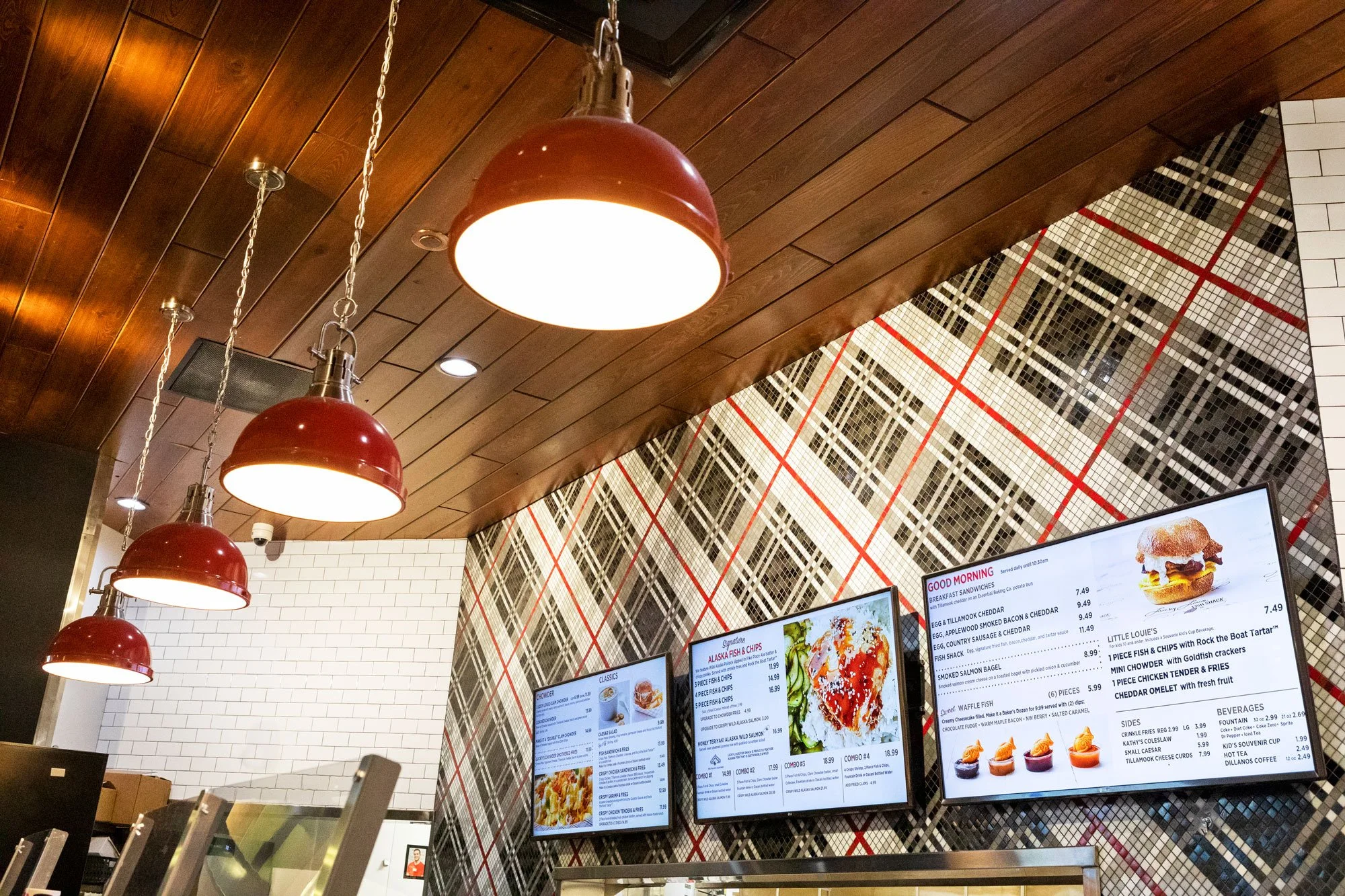 Interior of a restaurant or fast food establishment with a wooden ceiling, hanging red dome-shaped pendant lights, and a wall with white subway tiles and black and white checkered pattern. Multiple digital menu screens display food options and prices