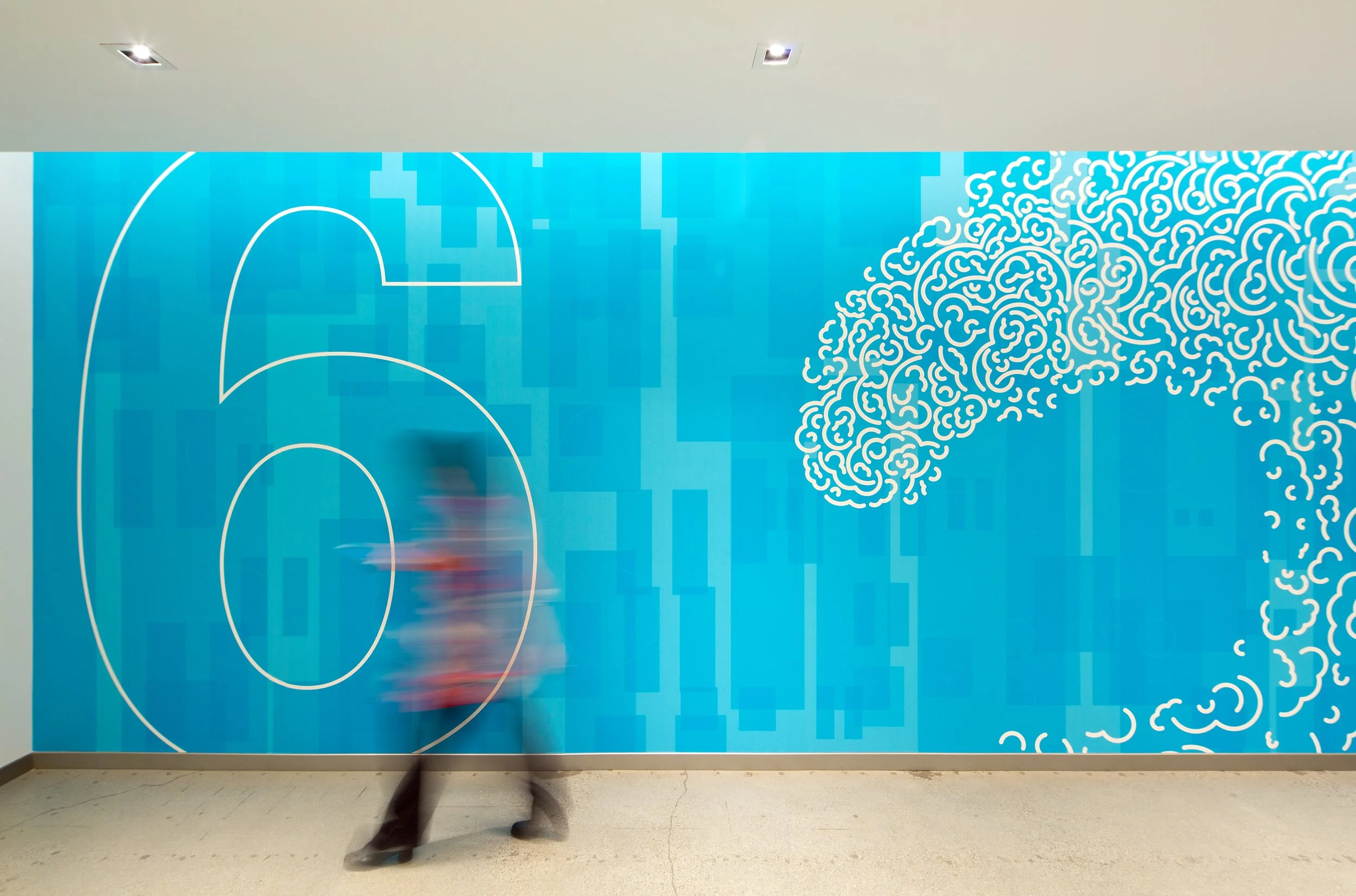 Large abstract blue wall mural with white line art design, including geometric shapes, concentric circles, and swirling patterns, in an indoor setting. Blurred figure walking by at the bottom.