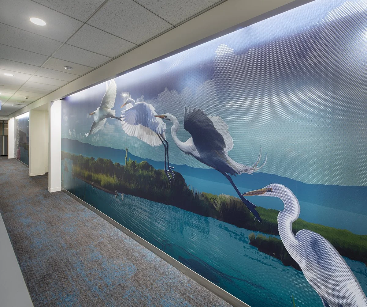 A mural of four herons flying over a river landscape with mountains and cloudy sky in the background, located on an indoor hallway wall.