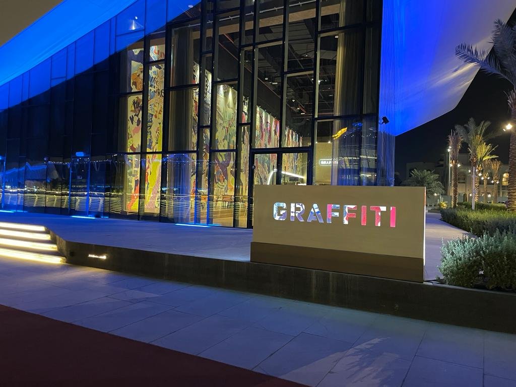Nighttime exterior of a modern building with glass walls, illuminated by blue and yellow lighting, featuring a large sign that reads "GRAFFITI" in front of the structure.