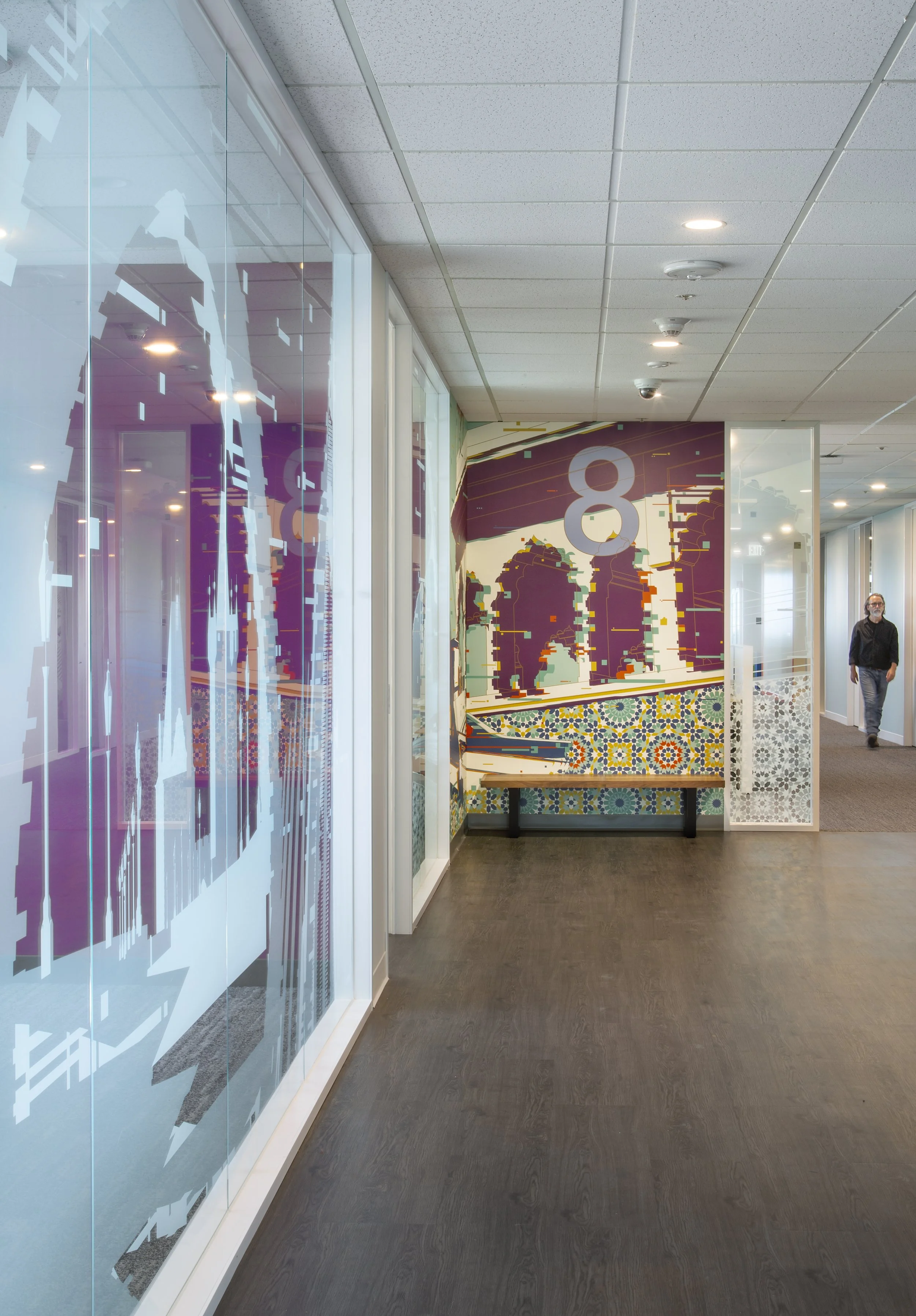 Modern office corridor with colorful mural and patterned tile artwork, glass walls, and a man walking.
