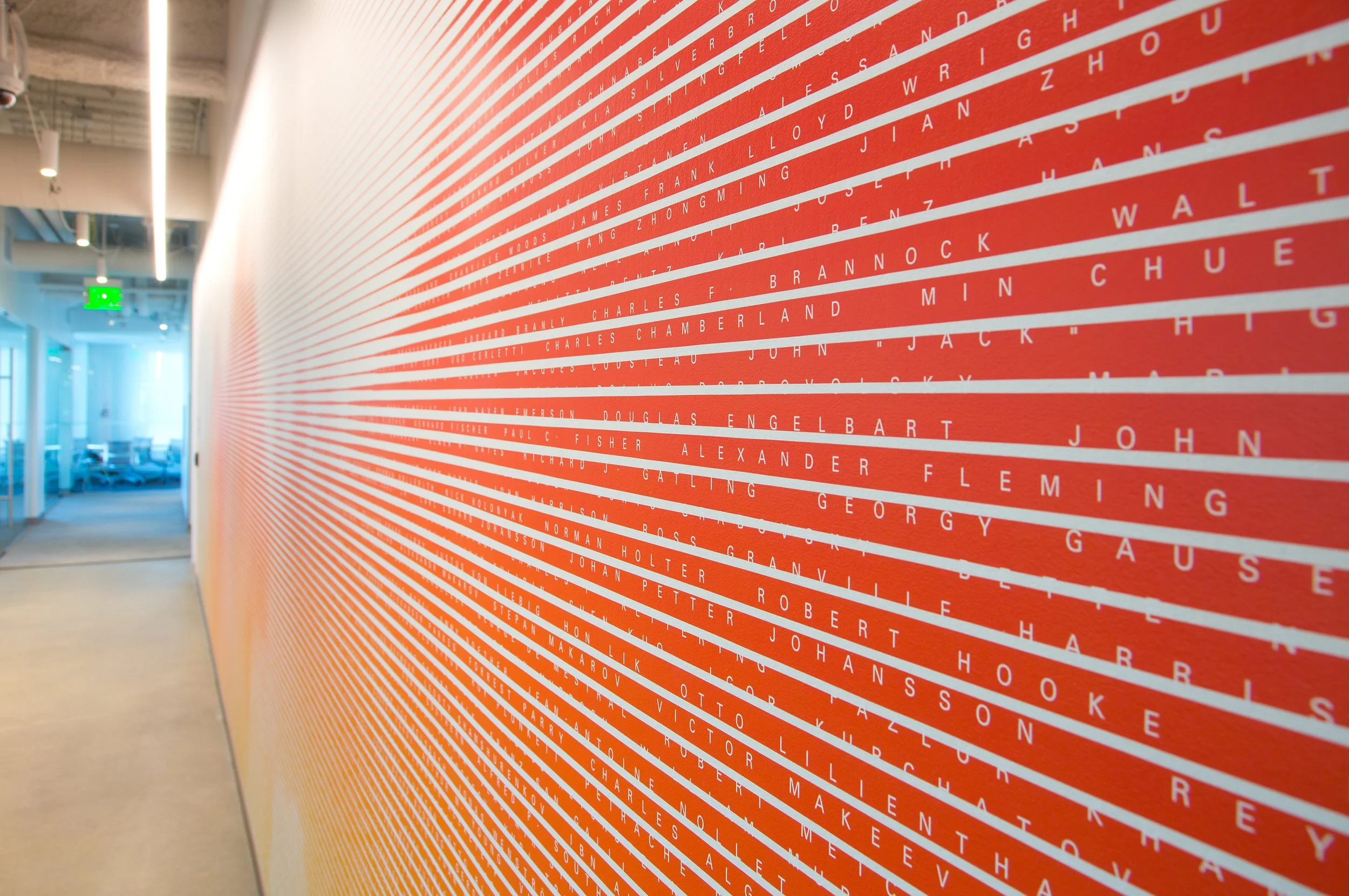 A large red wall with white diagonal lines and white text, listing names in a museum or memorial setting.