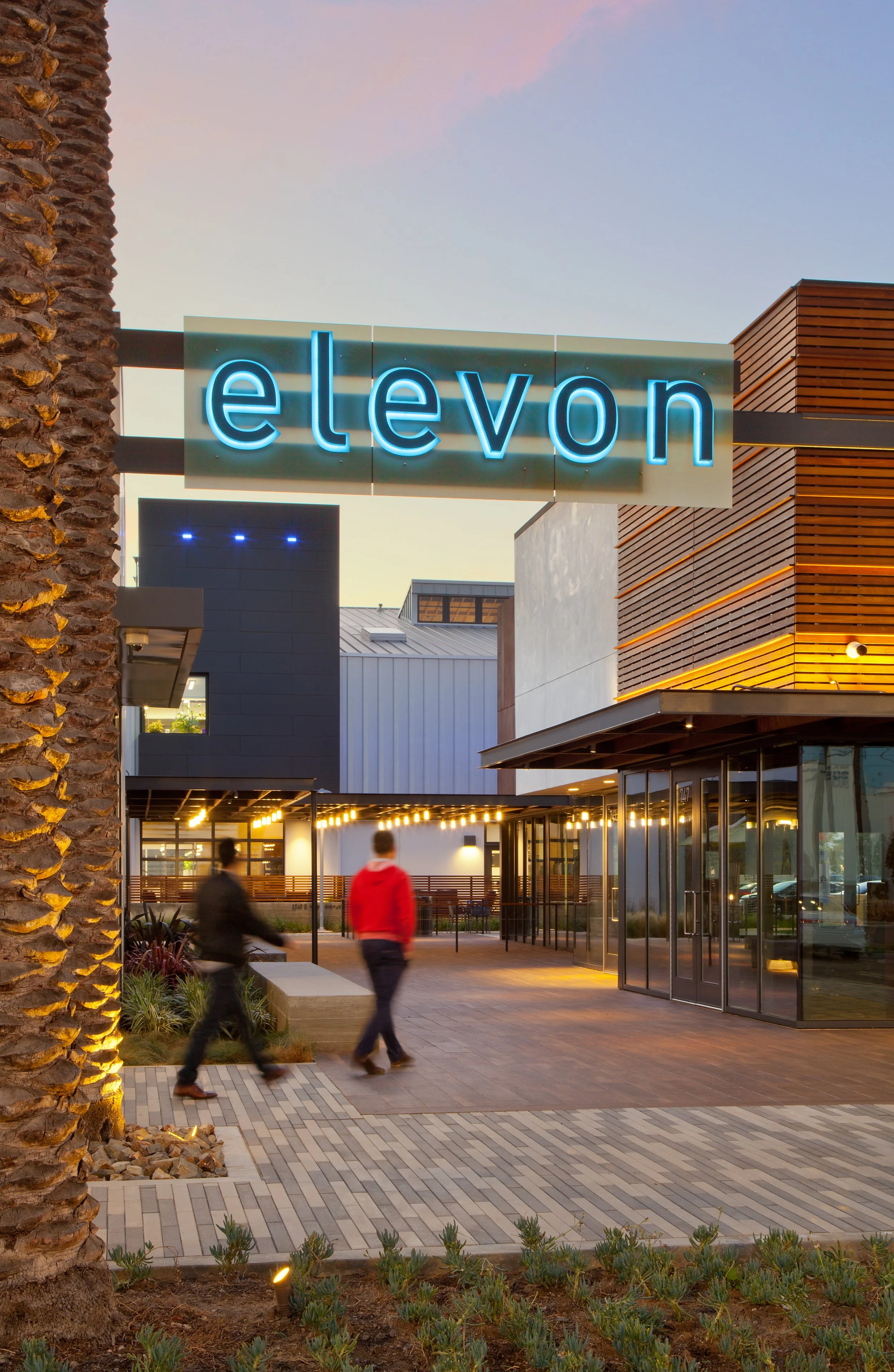 Exterior of a modern shopping or entertainment complex with a large illuminated sign reading 'eleven'. The building features wooden slats, glass windows, and a well-lit entrance. Two people are walking near the entrance, and there is landscaping with