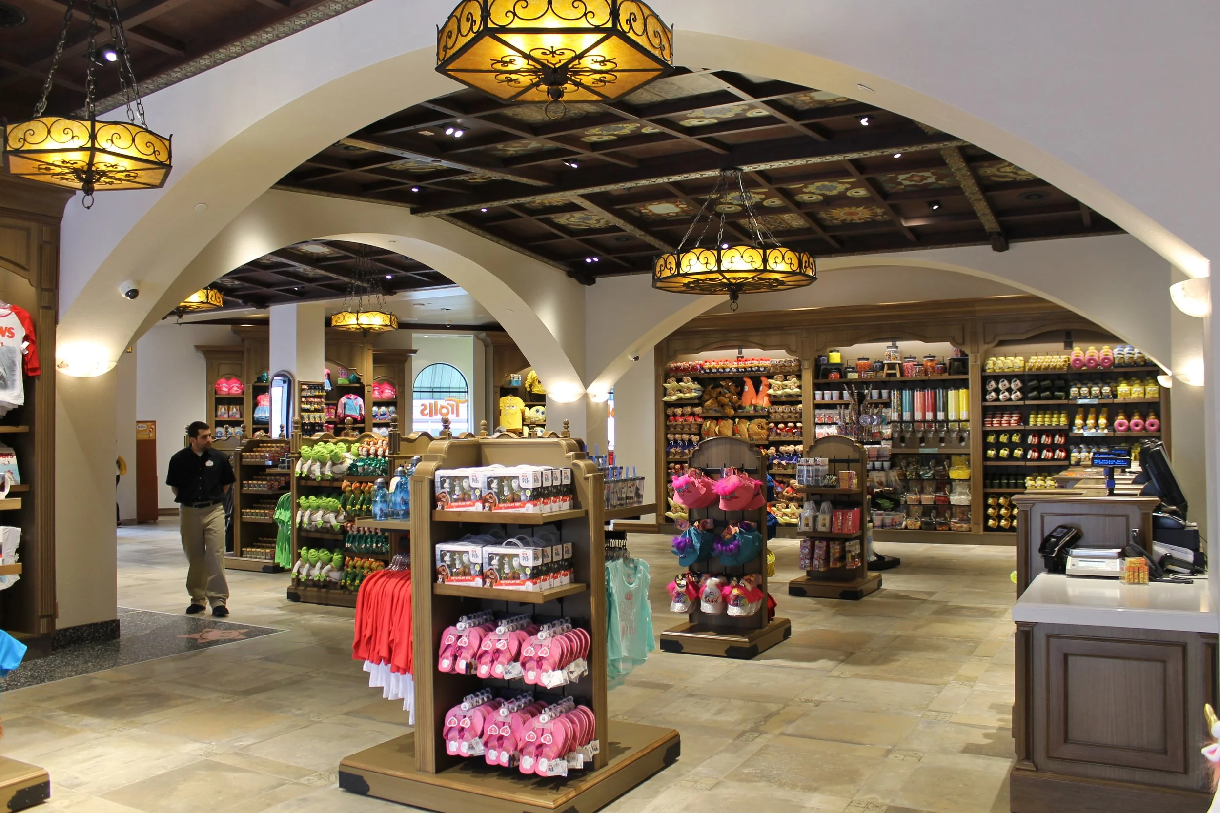 Inside a retail store with wooden shelves displaying toys, souvenirs, and merchandise. The ceiling features decorative wooden panels and hanging lights. A store employee is present near the entrance.