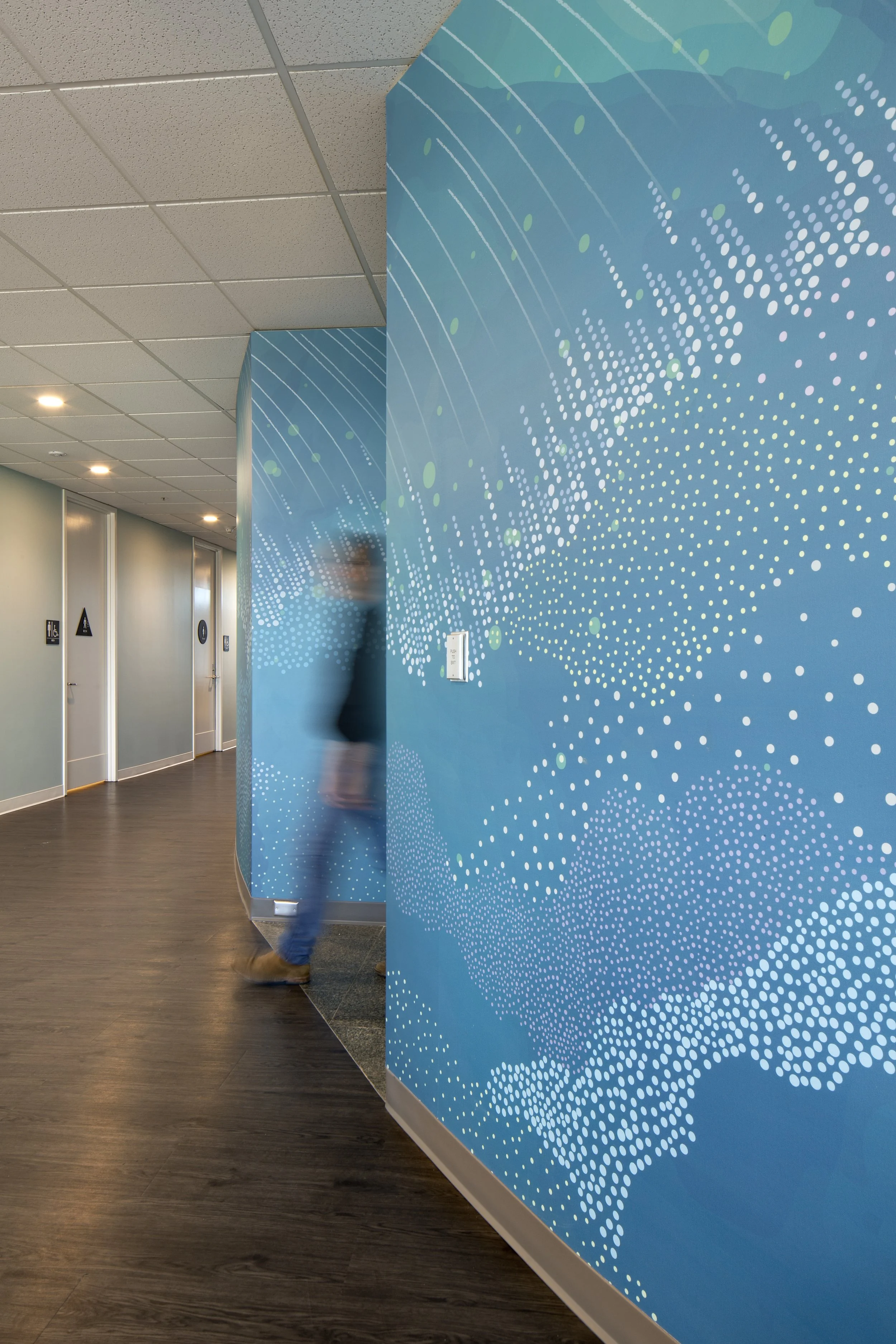 Indoor hallway with a blue wall featuring a dotted abstract pattern, beige ceiling tiles, and elevator doors in the background. A person is walking past the wall, slightly blurred.