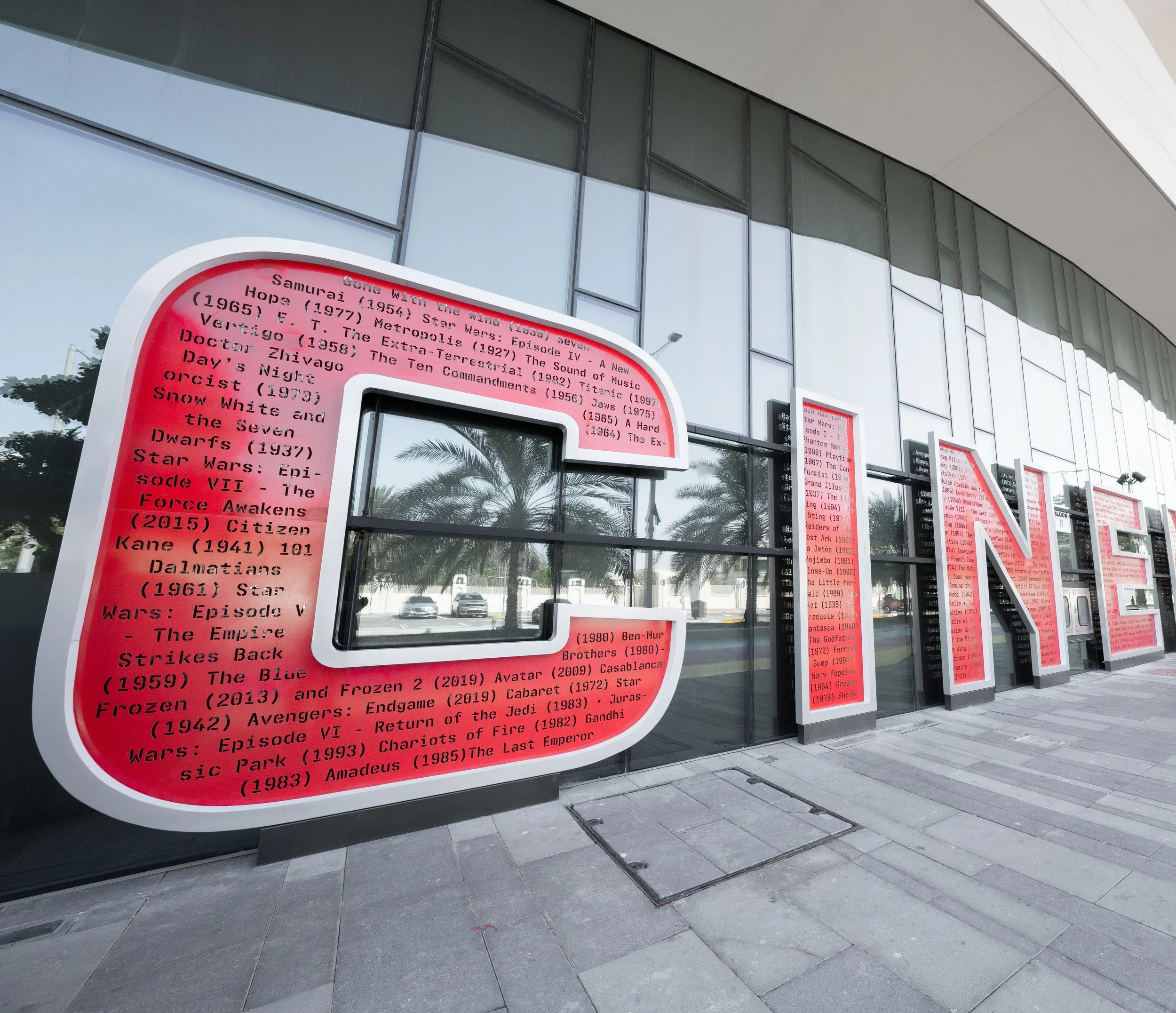 A modern building with glass windows and a curved white roof. Red and white electronic display boards are mounted on the exterior wall, listing titles of movies or television shows, with palm trees reflected in the glass windows.
