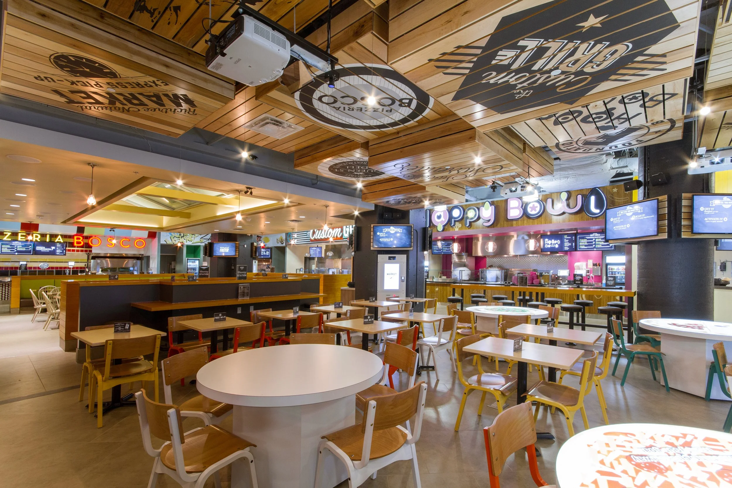 Inside a food court with various seating arrangements, digital menu boards, and restaurant signage including 'Happy Bowl' and 'Zeera Bosco'. The ceiling features wooden panels with logos and text, and there is a mounted projector.