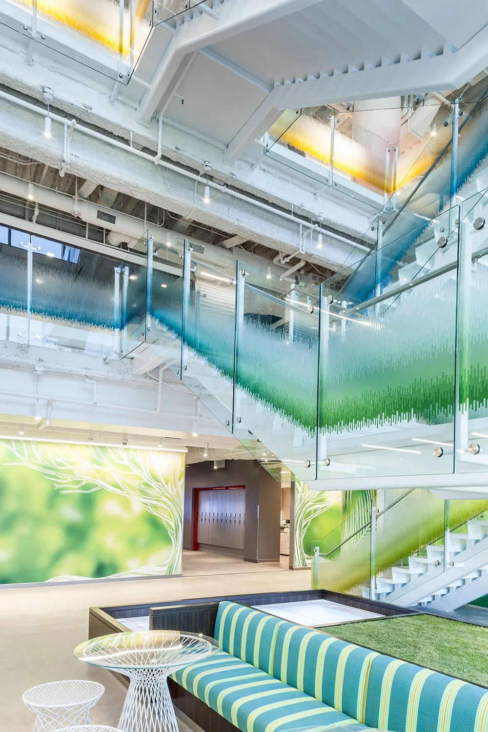 Modern office lobby with glass railings, colorful wall art depicting nature, and a seating area with striped blue and green cushions.