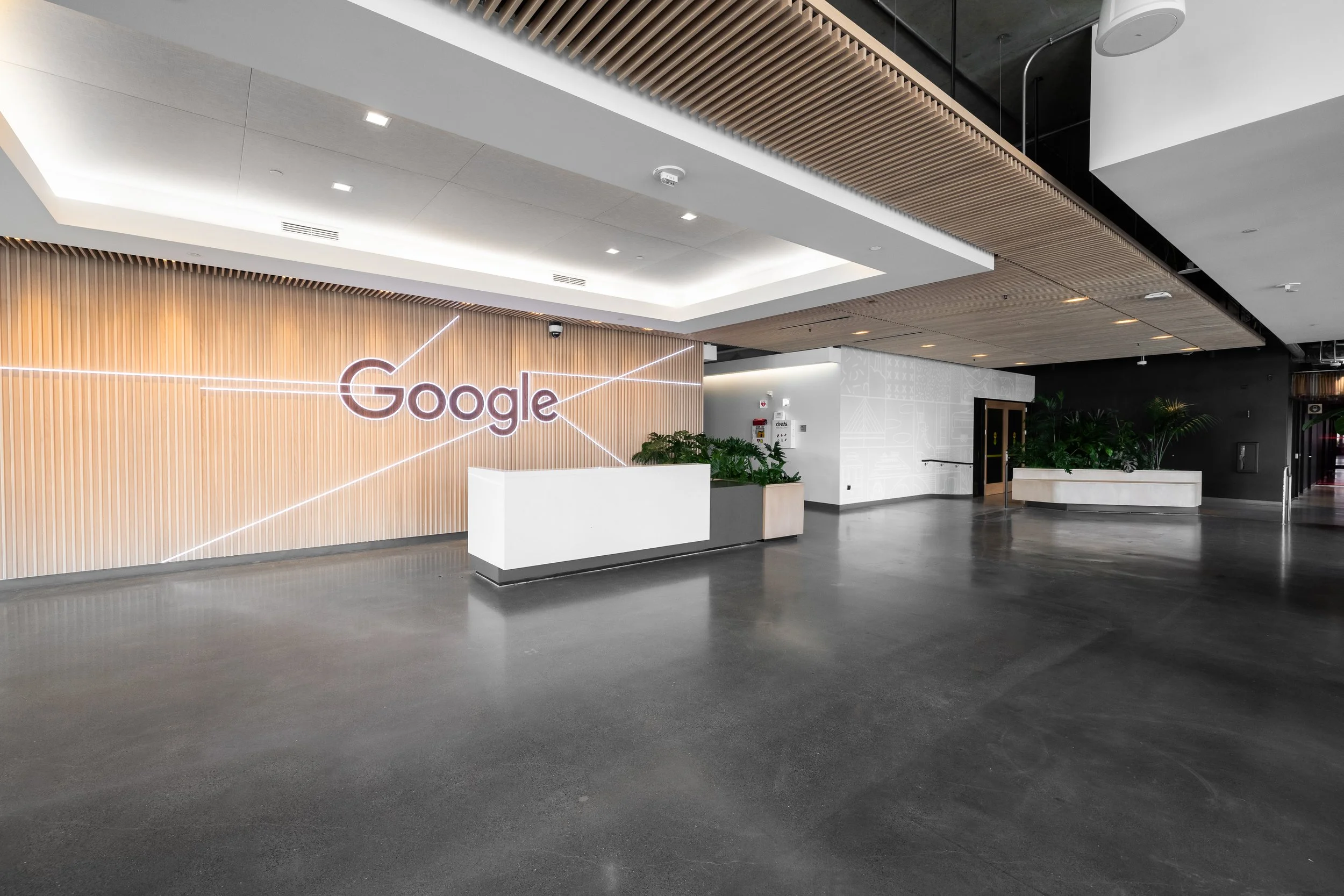 Interior view of a modern Google office reception area with a white reception desk, potted plants, and a large illuminated Google logo on a wooden-paneled wall.