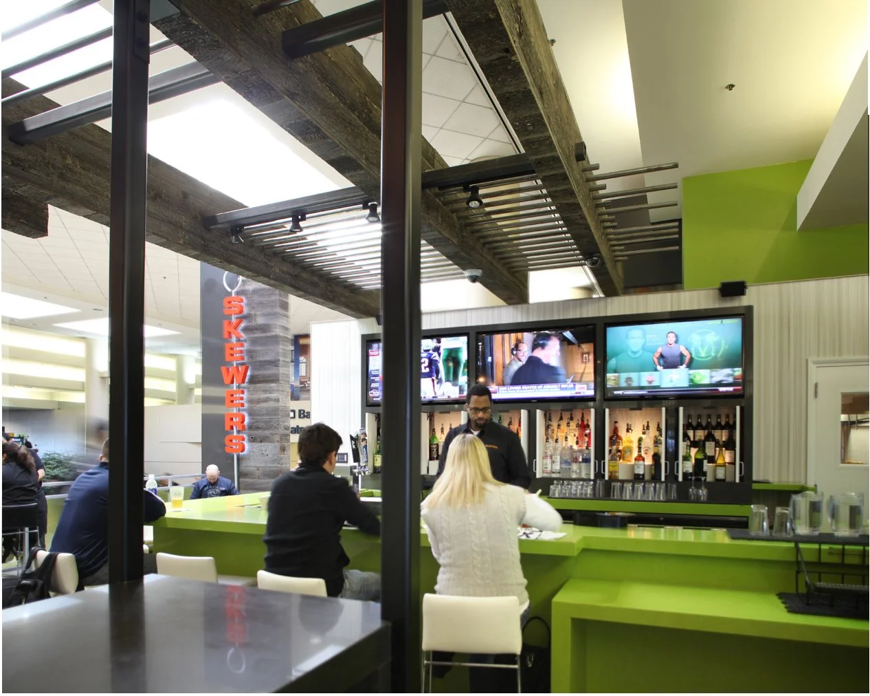 A bar area with a green counter, three people sitting, and a bartender behind the bar. Multiple television screens show news and sports, with a sign reading 'Bookers' on a concrete pillar. Other seating is visible in the background.