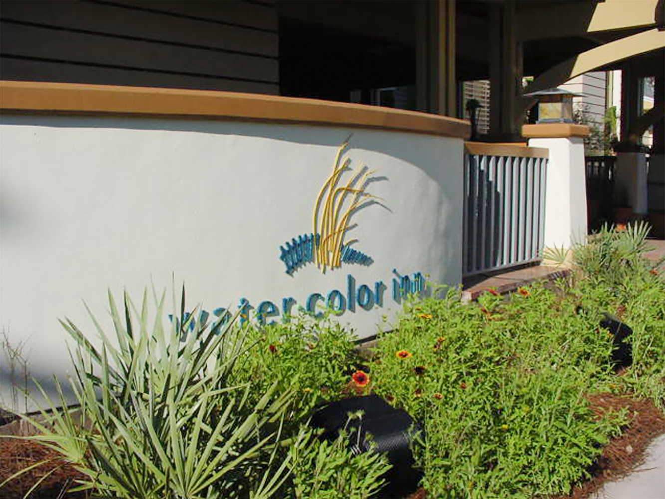 Exterior view of the entrance to a building with a white wall, a sign that reads 'watercolor inn' with a graphic of wheat and grass, surrounding plants, and part of a porch with a gray railing.