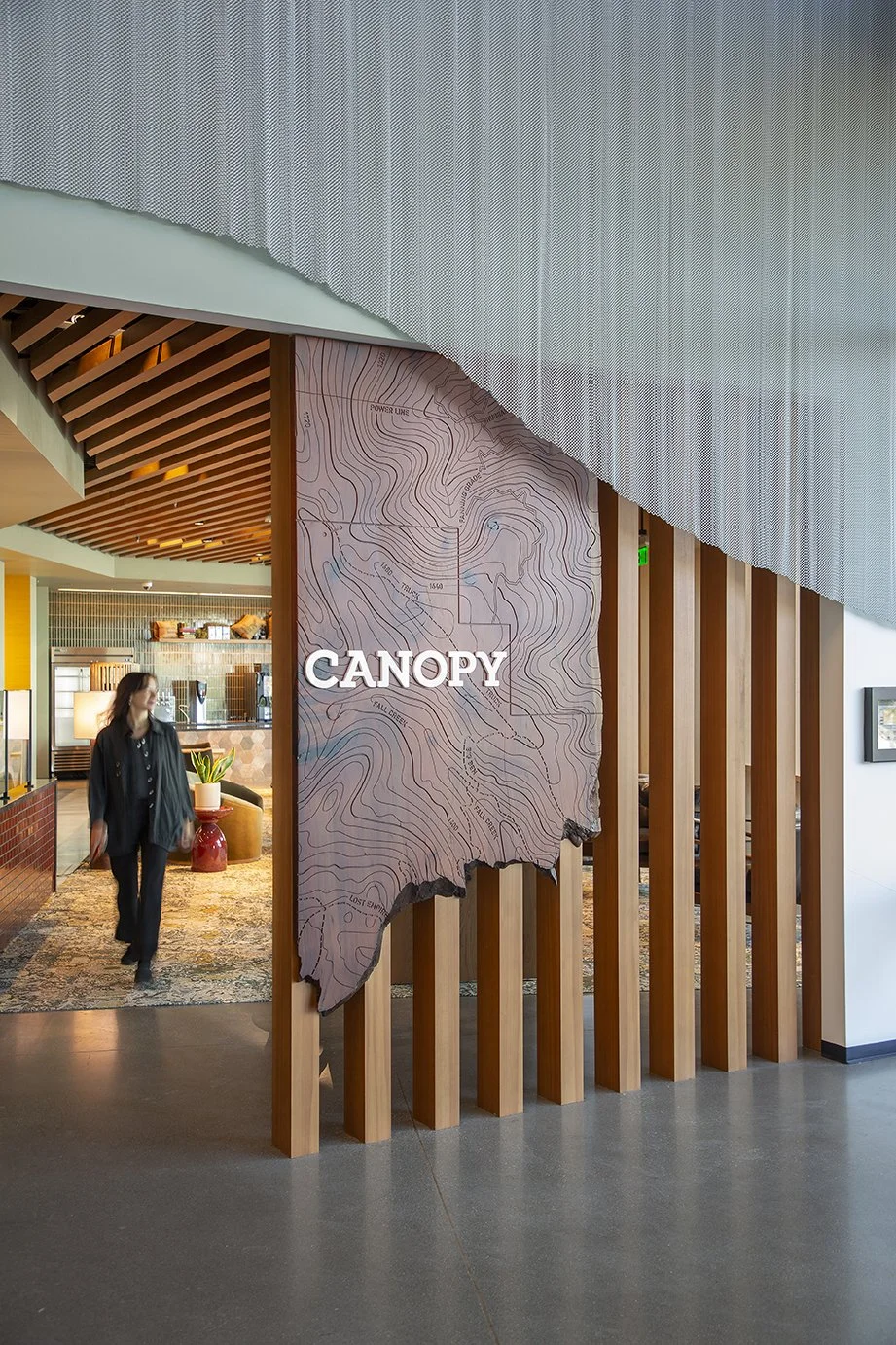 Interior of a building with a sign that says 'CANOPY' on a wooden and topographical map decor, featuring warm lighting, wooden vertical slats, a reception area with a woman walking by, and modern design elements.