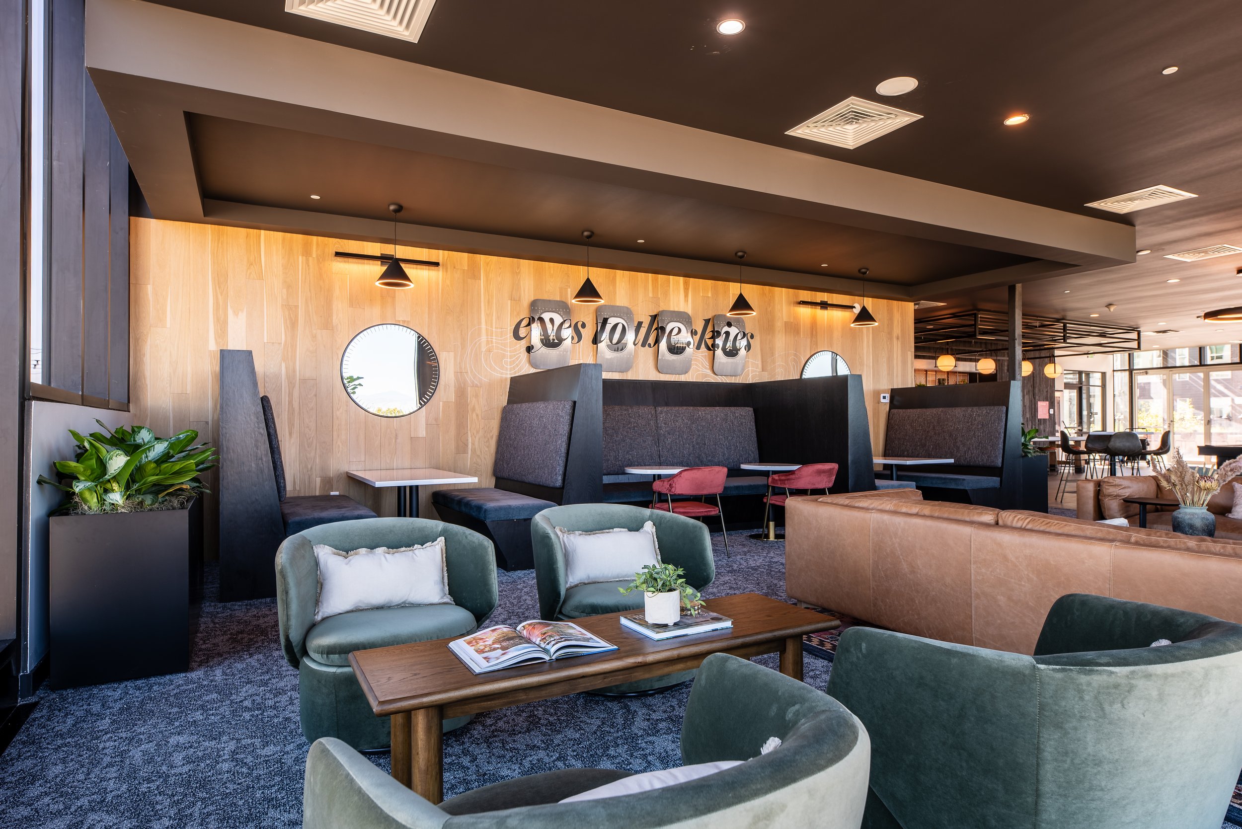 Modern hotel lobby with seating area, including green and gray chairs, a wooden coffee table with open magazines and a small plant, a leather sofa, a wooden wall with circular windows, pendant lights, and a sign that reads 'eyes to the kin' on the wa