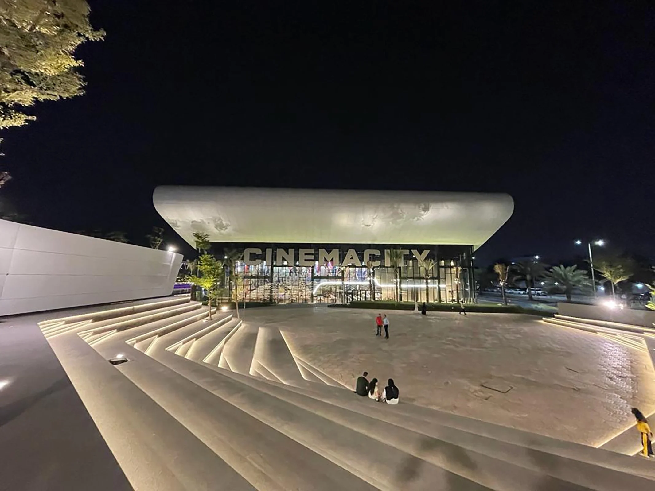 Night view of the exterior of a modern building with large 'CINEMATEX' sign and unique architectural stairs leading up to it. There are small groups of people sitting on the steps, and trees are visible near the building.