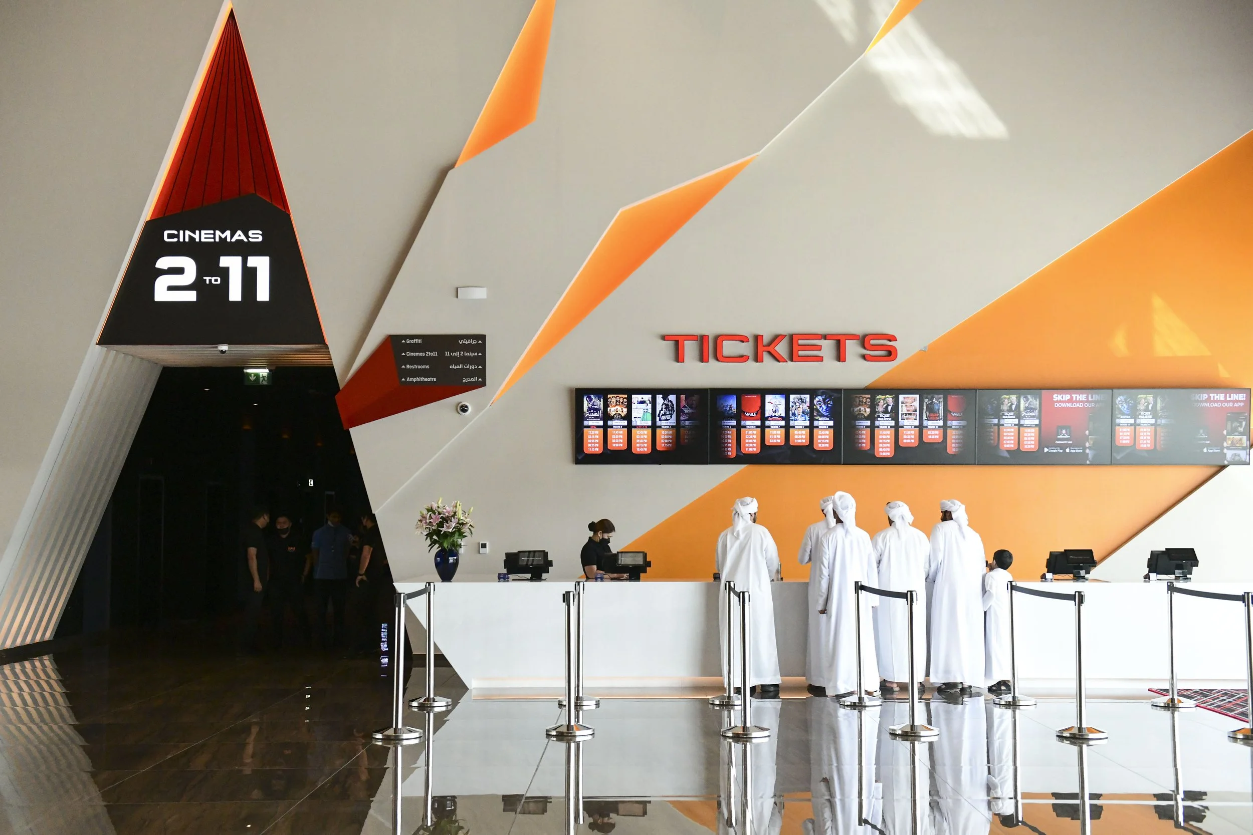A modern movie theater ticket counter with staff and guests, a digital display of movie posters and showtimes, and a sign indicating ticket sales from 2 to 11. Decorative orange and gray geometric wall panels.