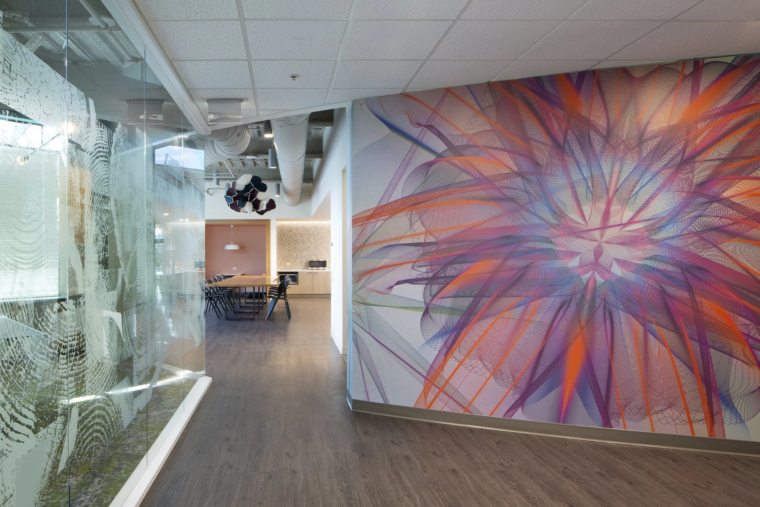 Modern office interior with a colorful abstract mural on one wall, glass partition with etched fingerprints, and a conference table with chairs. Ceiling has exposed ducts and hanging light fixtures.