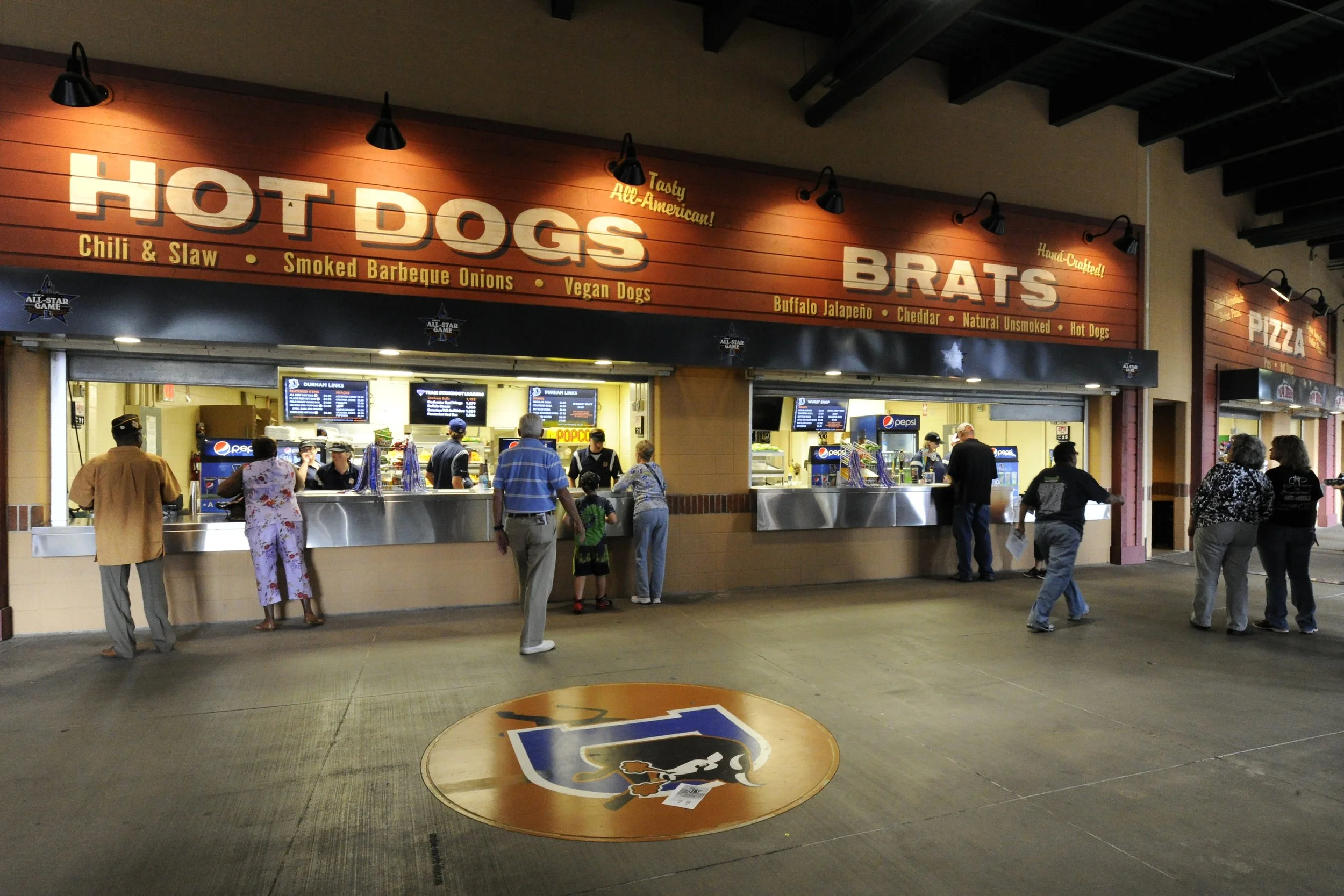 A food stand with signs reading HOT DOGS and BRATS, serving chili and slaw, smoked barbeque onions, vegan dogs, buffalo jalapeño, cheddar, natural unsmoked, and hot dogs. Several people are standing in line or ordering, with digital menu screens visi