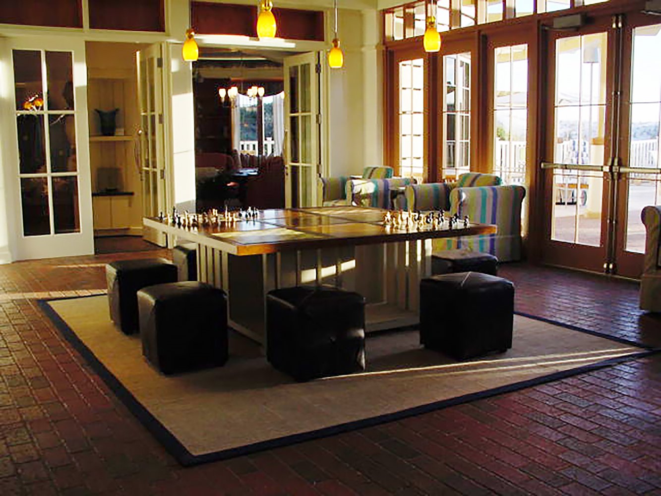 Interior of a sunlit room with a chess table at the center, surrounded by black ottomans. Large windows and glass doors let in natural light, with colorful striped chairs and wooden accents.