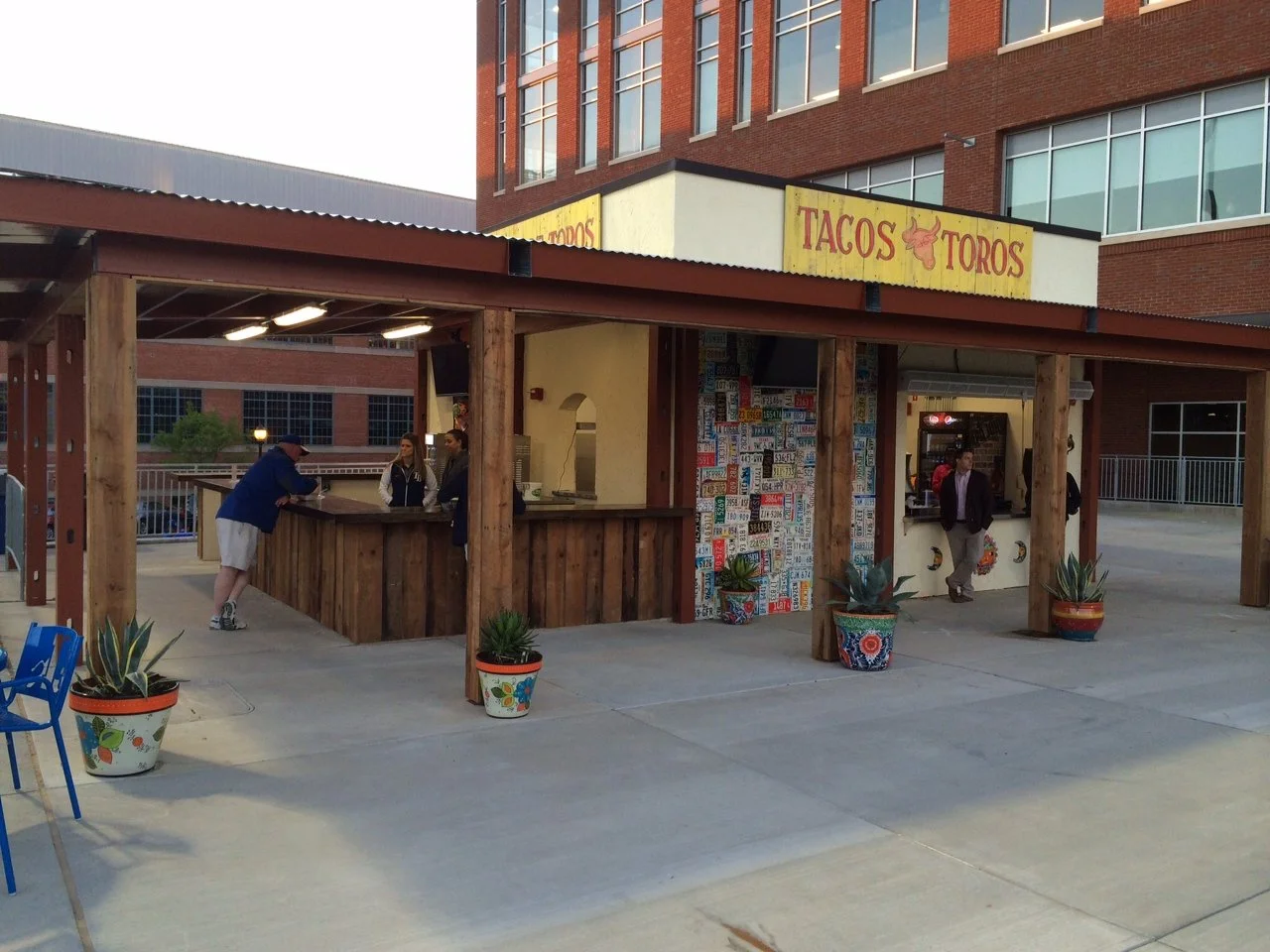 Outdoor taco stand with a yellow sign that reads Tacos Toros and has a bull head logo. The stand has a wooden counter, potted plants, and license plates on the wall. Two people are ordering and two staff members are working.