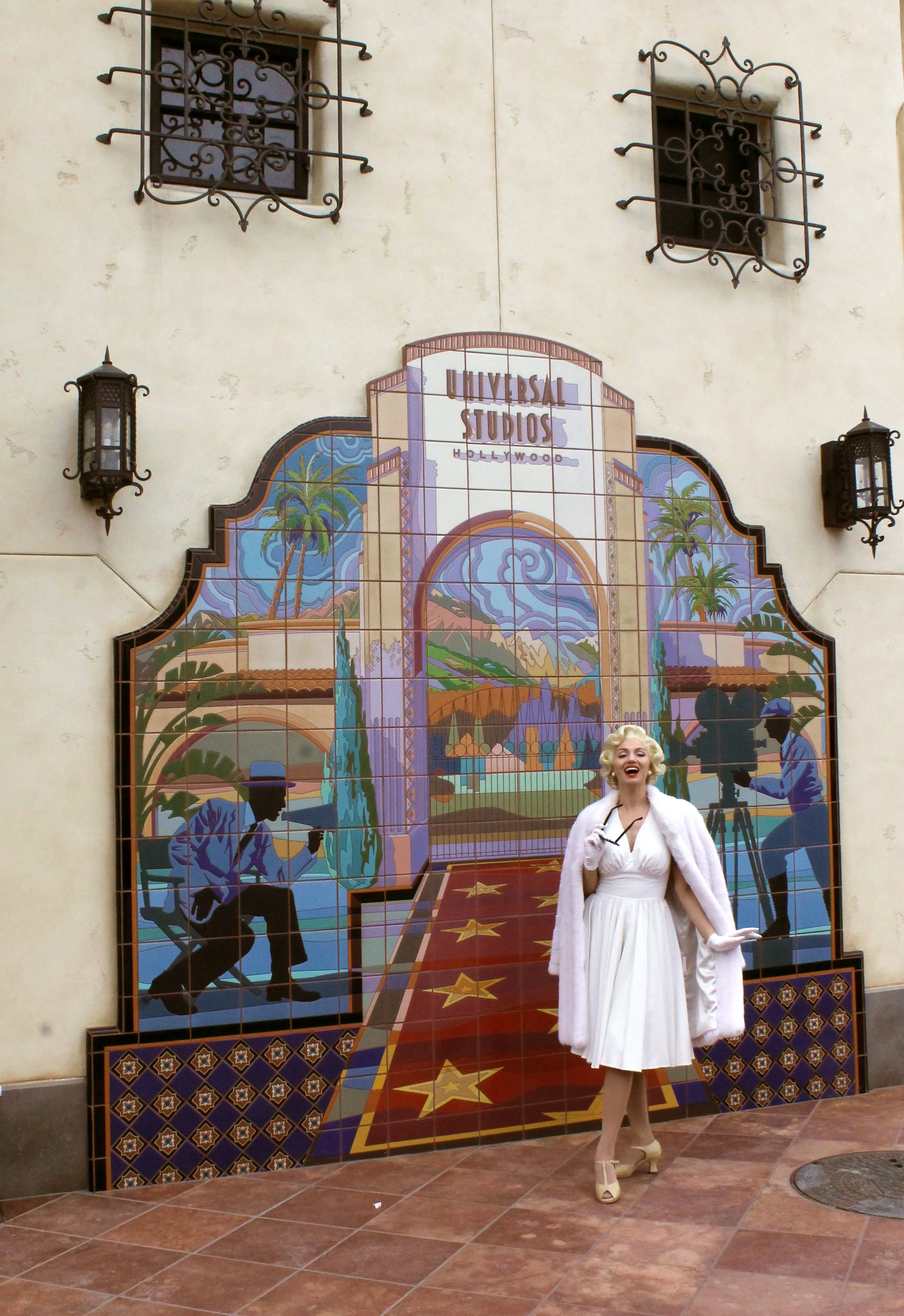 A woman dressed as Marilyn Monroe standing in front of a mural of the Hollywood sign at Universal Studios. She is wearing a white dress, a pink fur coat, and beige high heels.