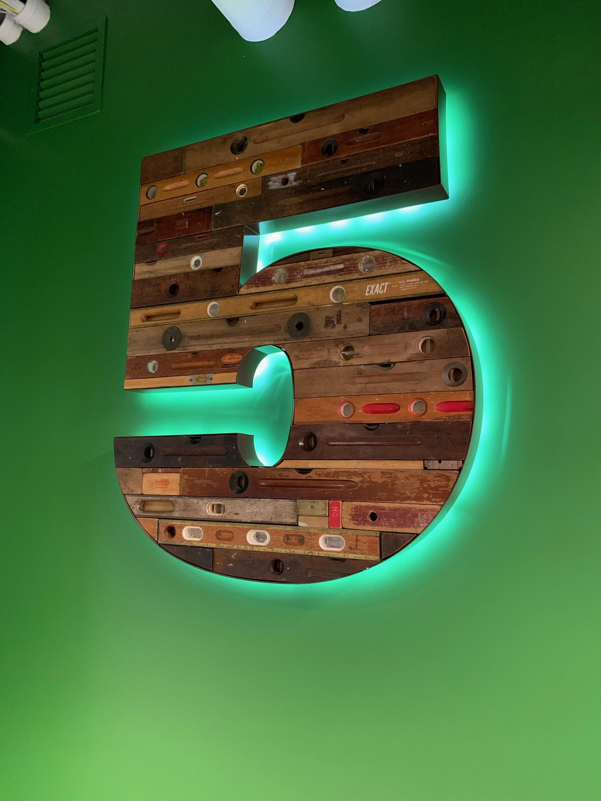 A large number '5' made of wooden planks with illuminated green backlighting on a green wall.