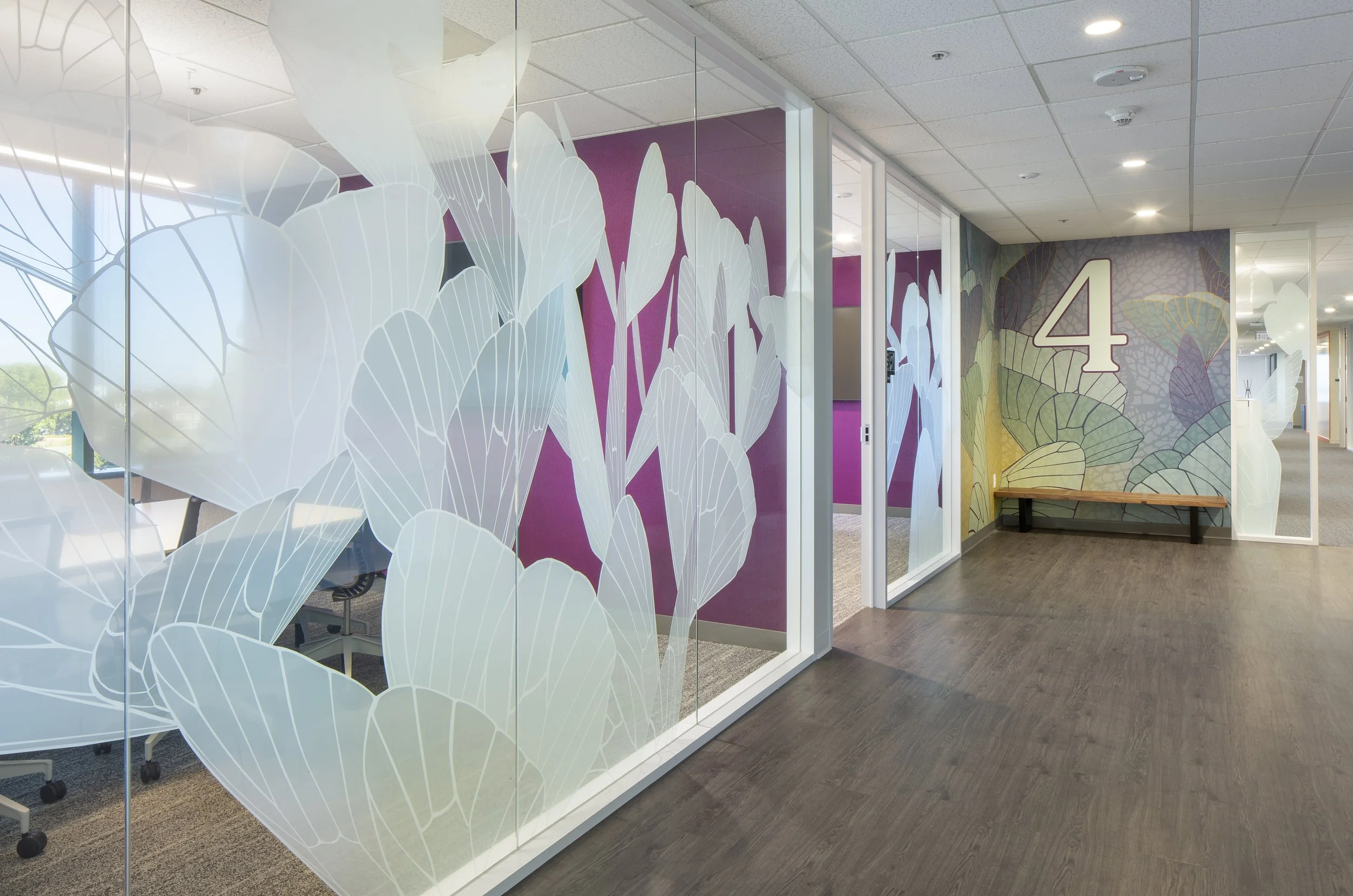 Office hallway with glass partitions decorated with white leaf patterns, a large number 4 on the wall, colorful mural, and wooden floor.