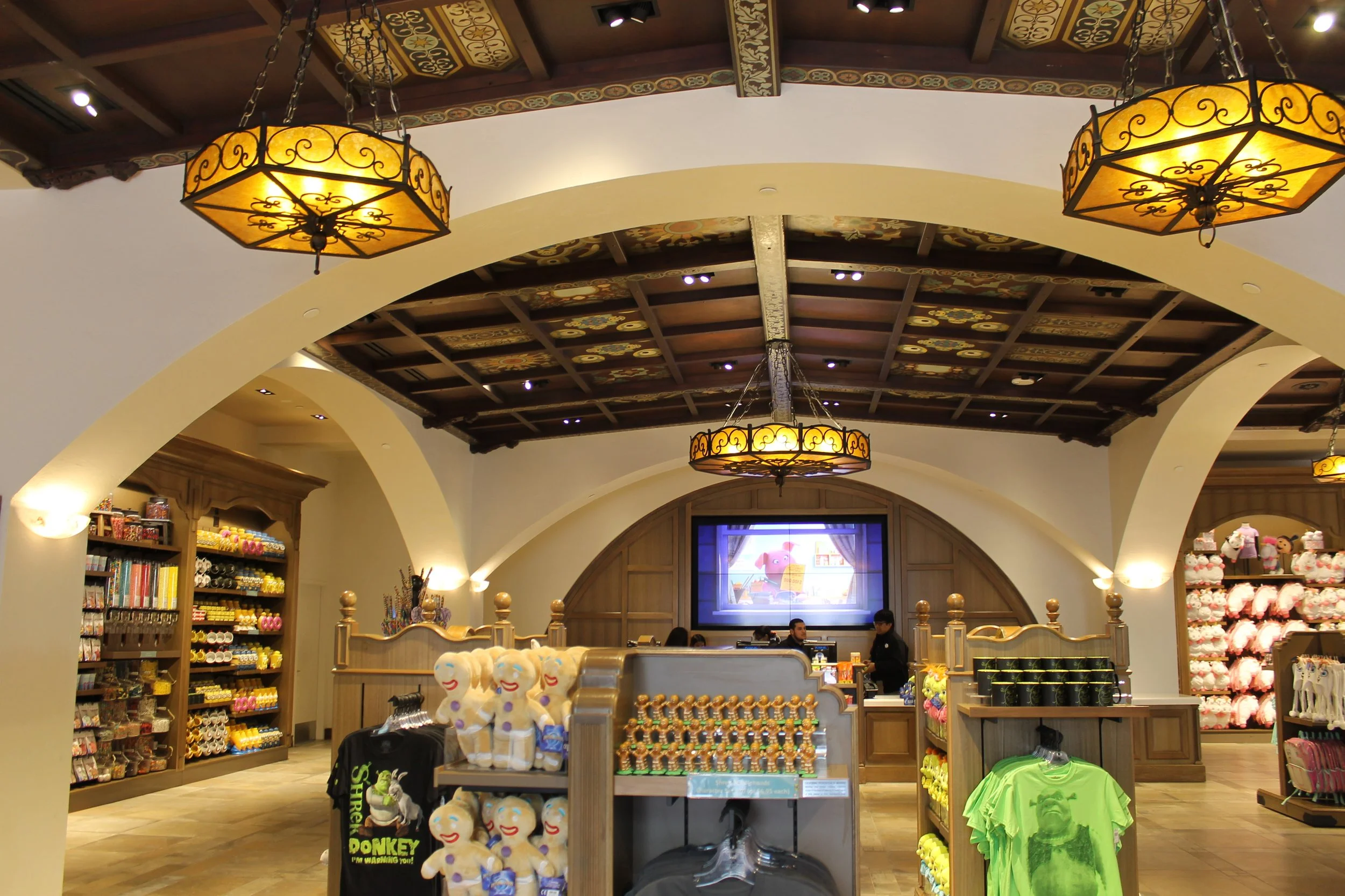 Inside a retail store with wooden display shelves, plush toys, and colorful merchandise, featuring ornate ceiling chandeliers, decorative wooden beams, and a large monitor screen showing animated characters.