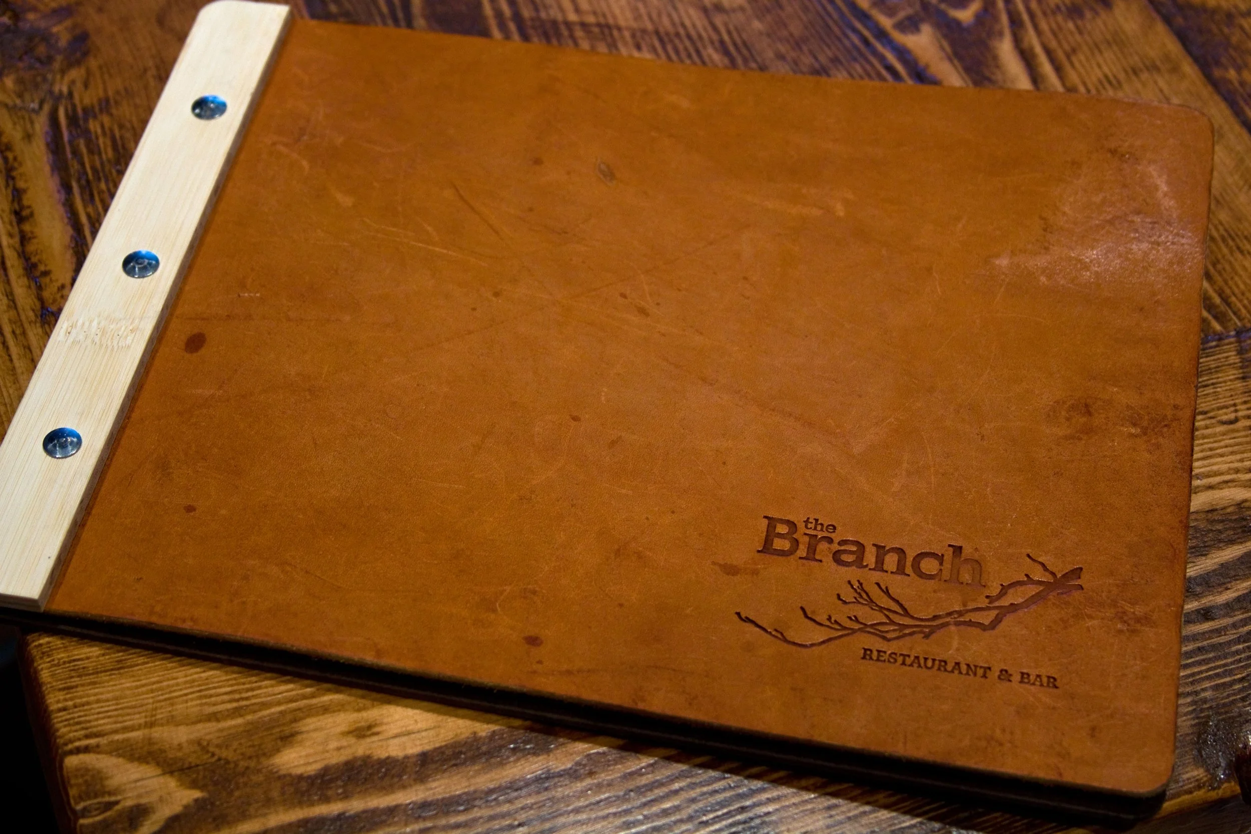 A brown paper menu with the words 'the Branch' and a tree branch graphic, placed on a wooden table.