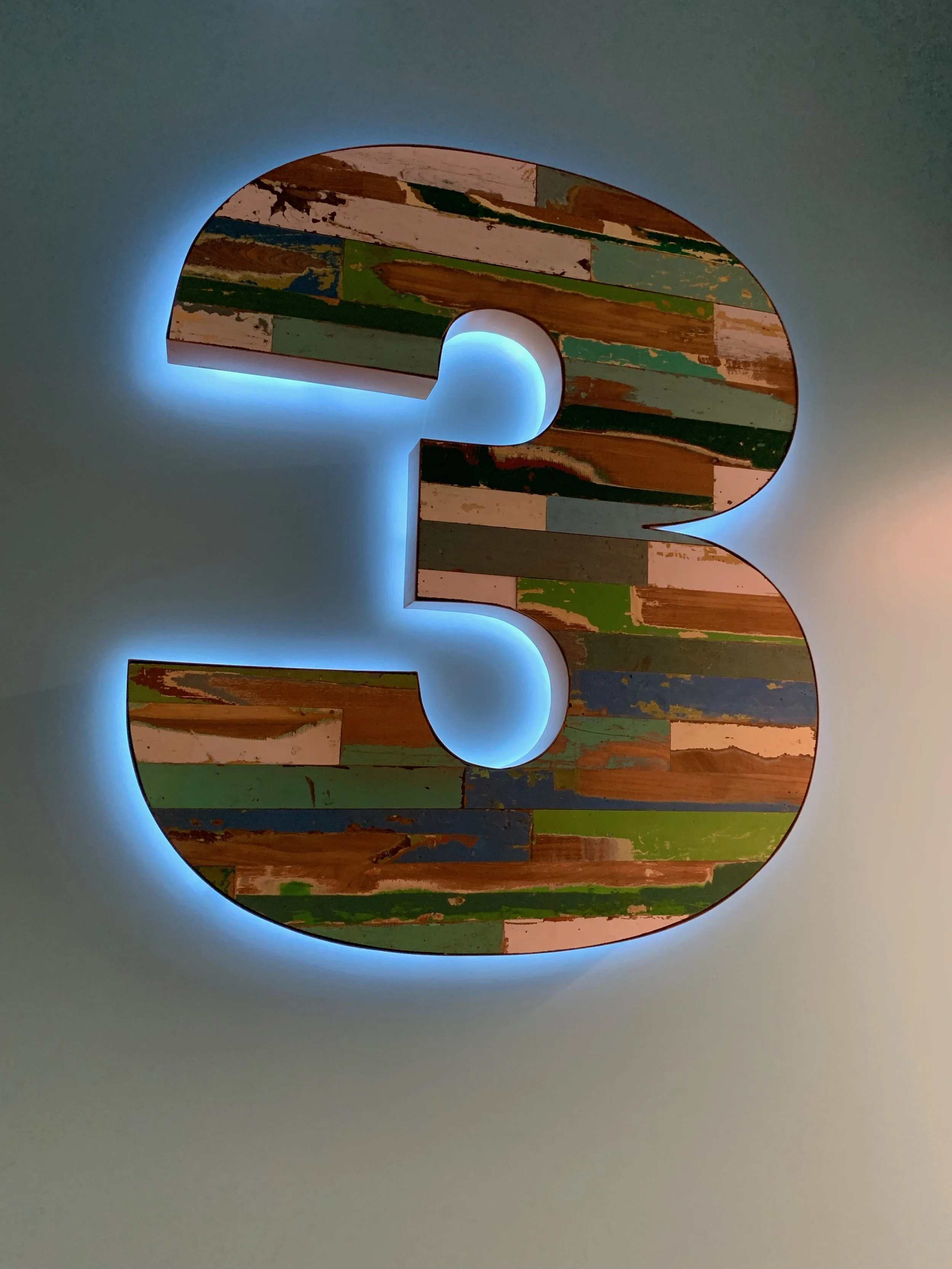 Decorative number 3 with colorful distressed wood pattern and backlit blue glow on a gray wall.