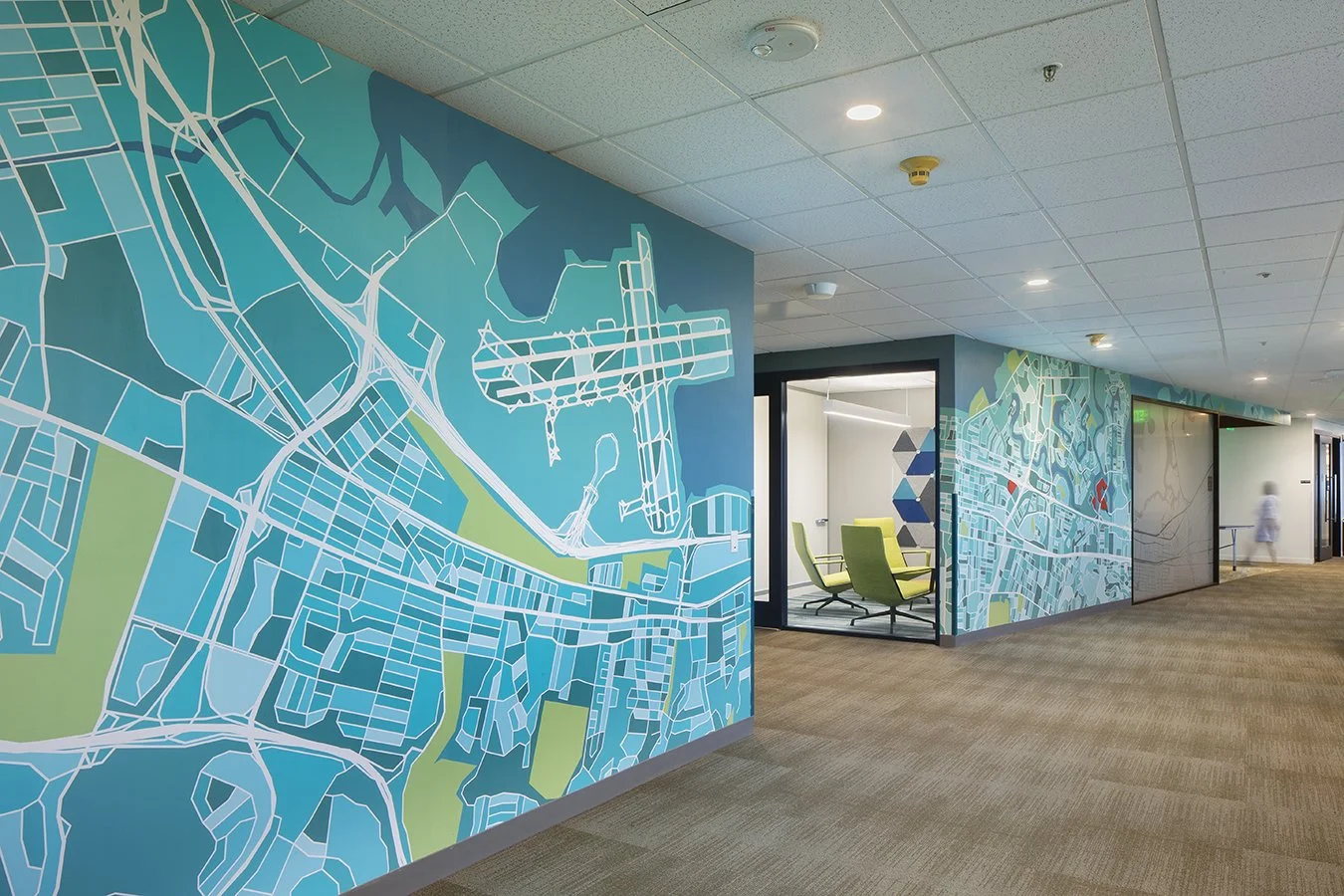 Office hallway with colorful wall murals resembling city maps, glass-walled meeting rooms, and people walking in the distance.