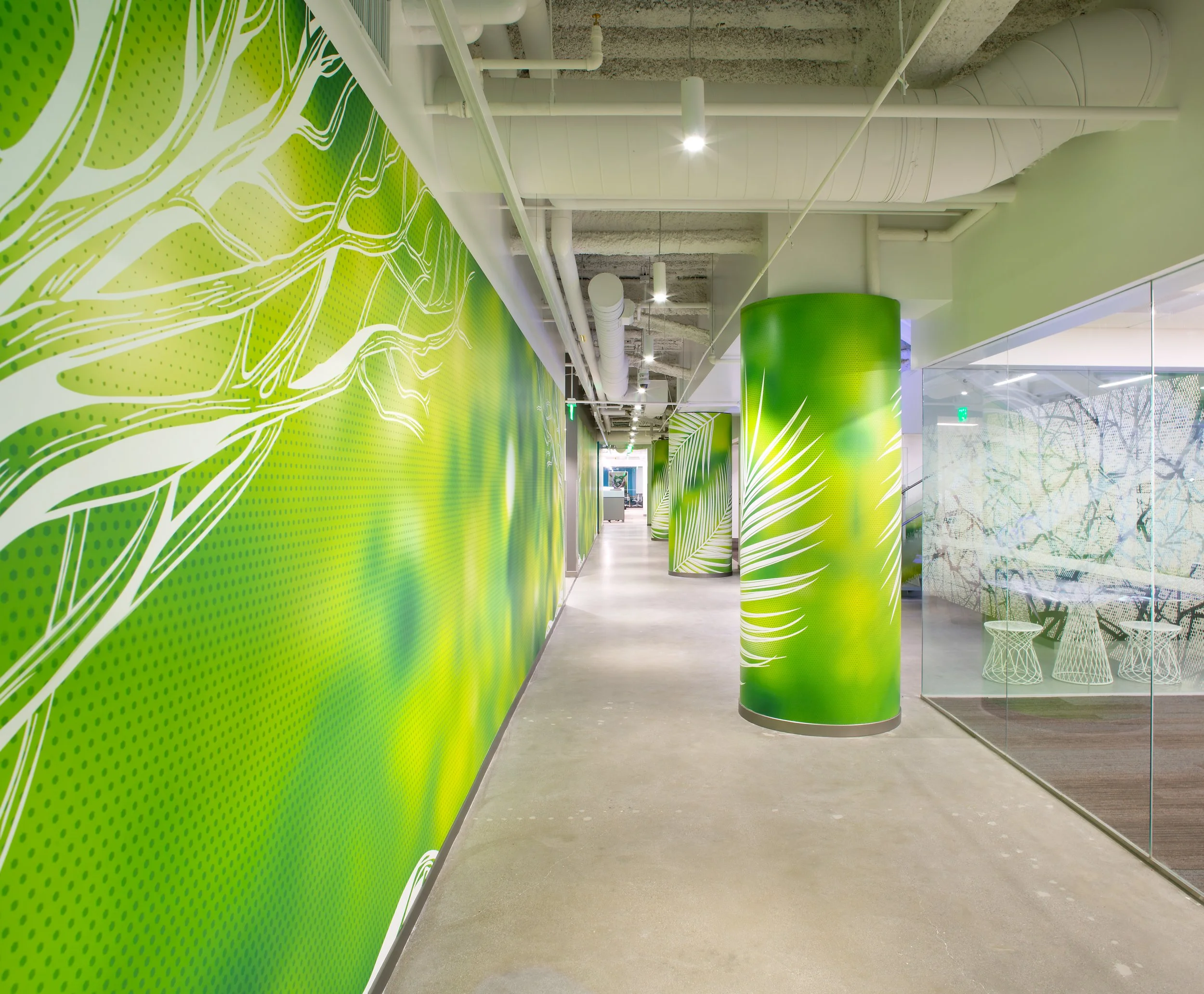 Modern office corridor with green and white decorative wall art, green cylindrical pillars, exposed ductwork, and concrete ceiling.