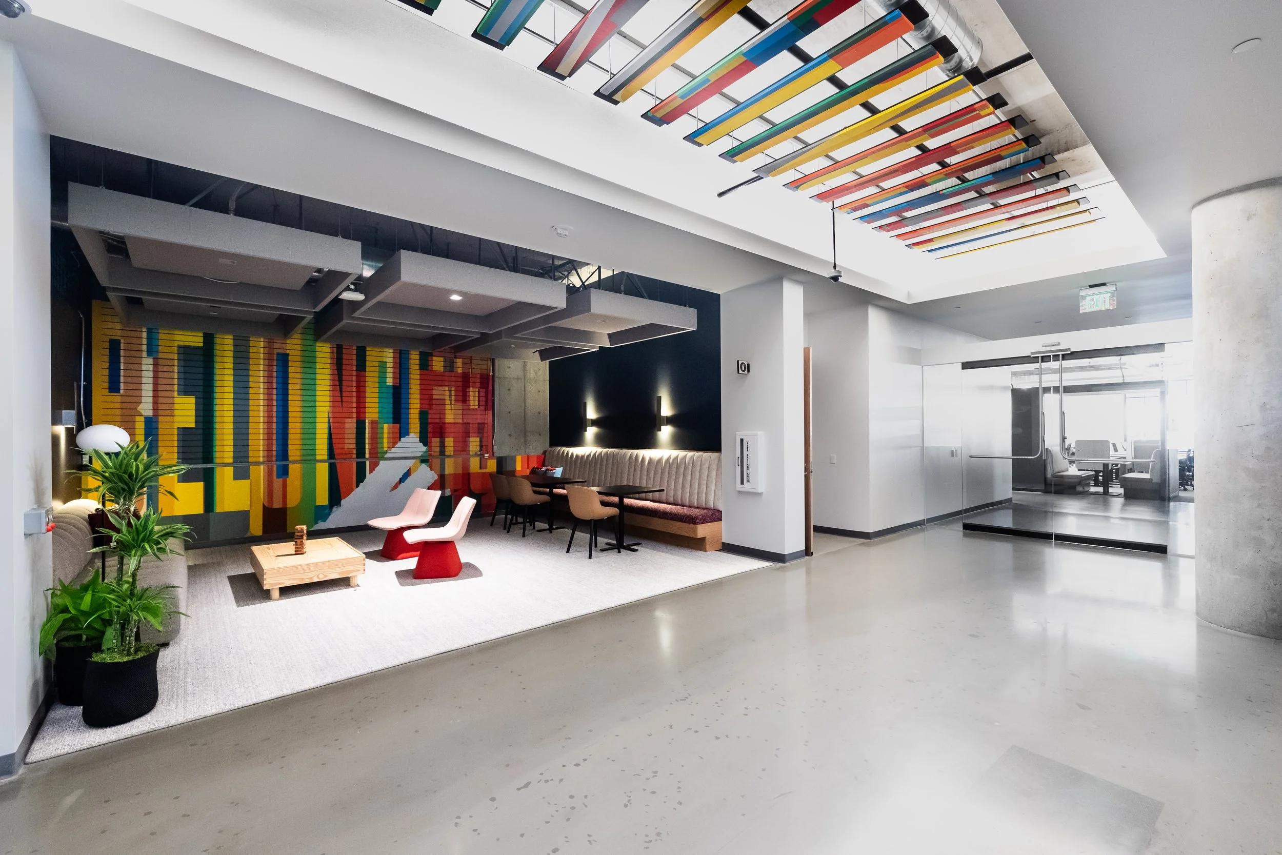 Modern hotel lobby with colorful ceiling art, seating area with beige and pink chairs, small wooden table, potted plants, and a vibrant wall mural. Glass doors lead to another room or outdoor area.