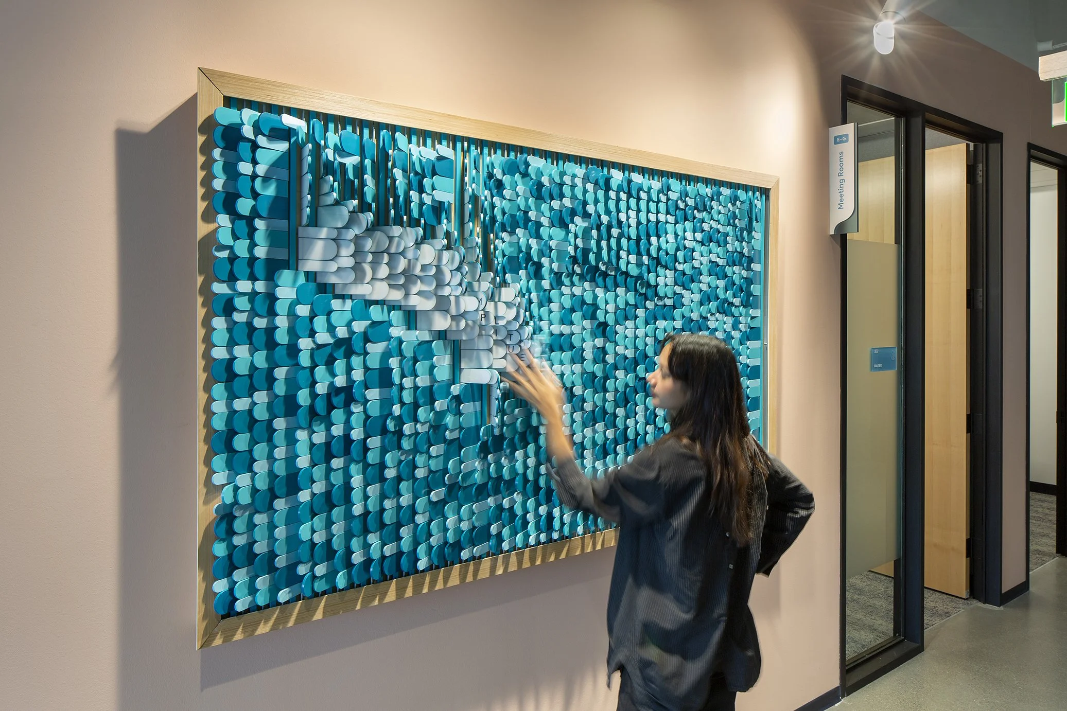 Woman arranging a blue and white textured wall art piece in an office hallway.